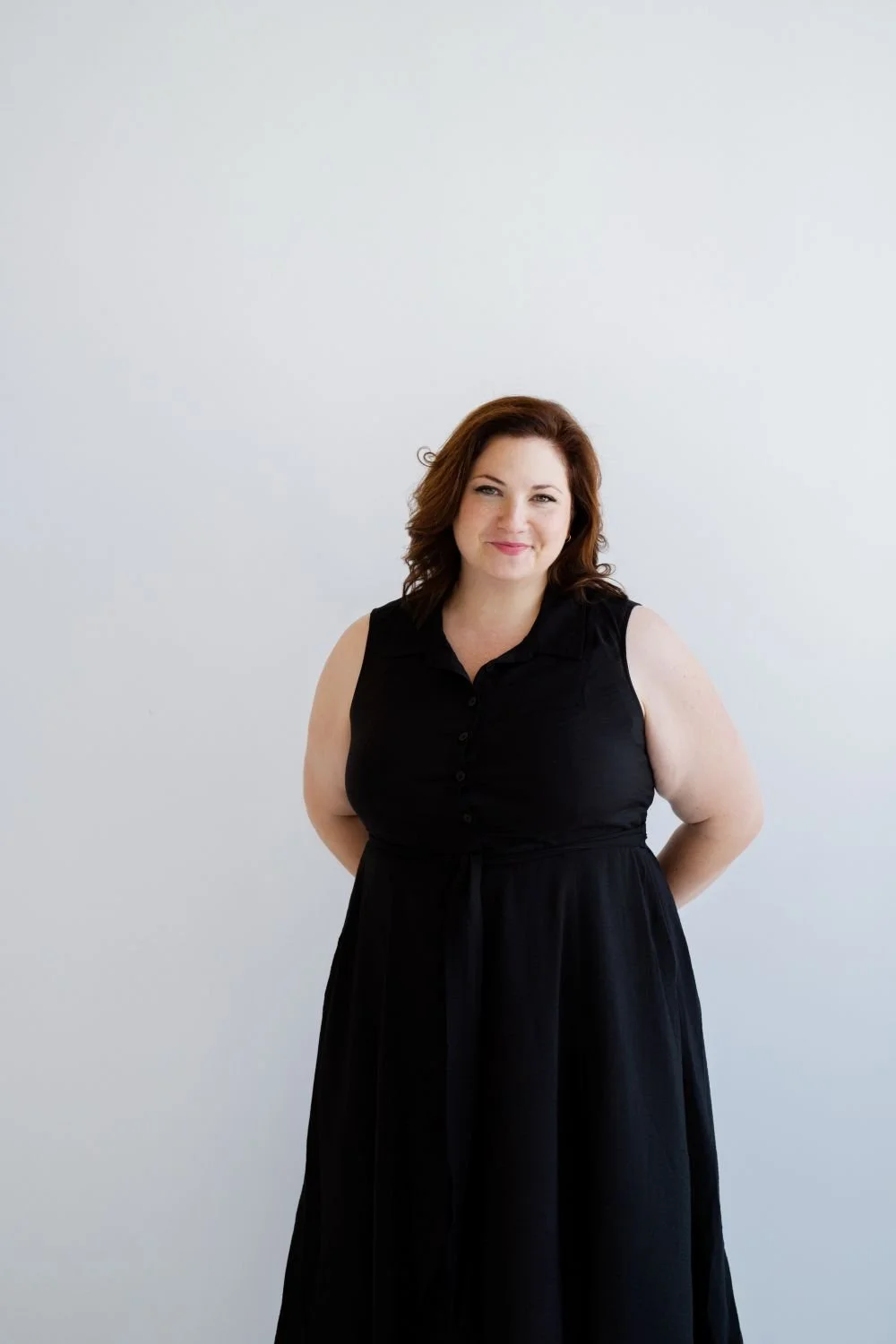 A woman with shoulder-length brown hair wearing a sleeveless black dress standing against a plain white background.