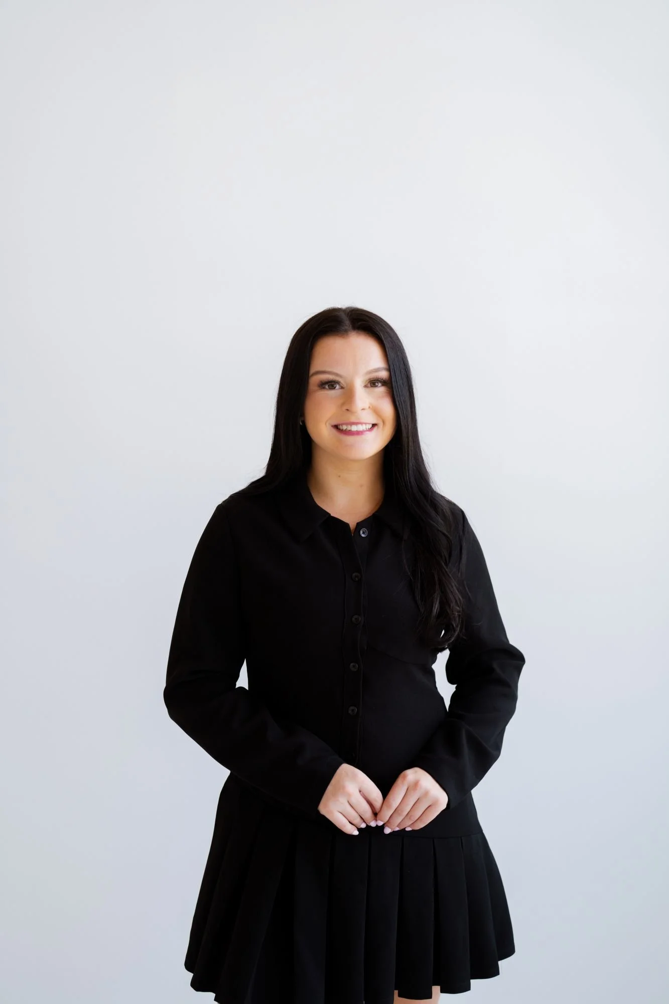 A woman with long dark hair, wearing a black button-up shirt and black skirt, standing against a plain white wall, smiling at the camera.