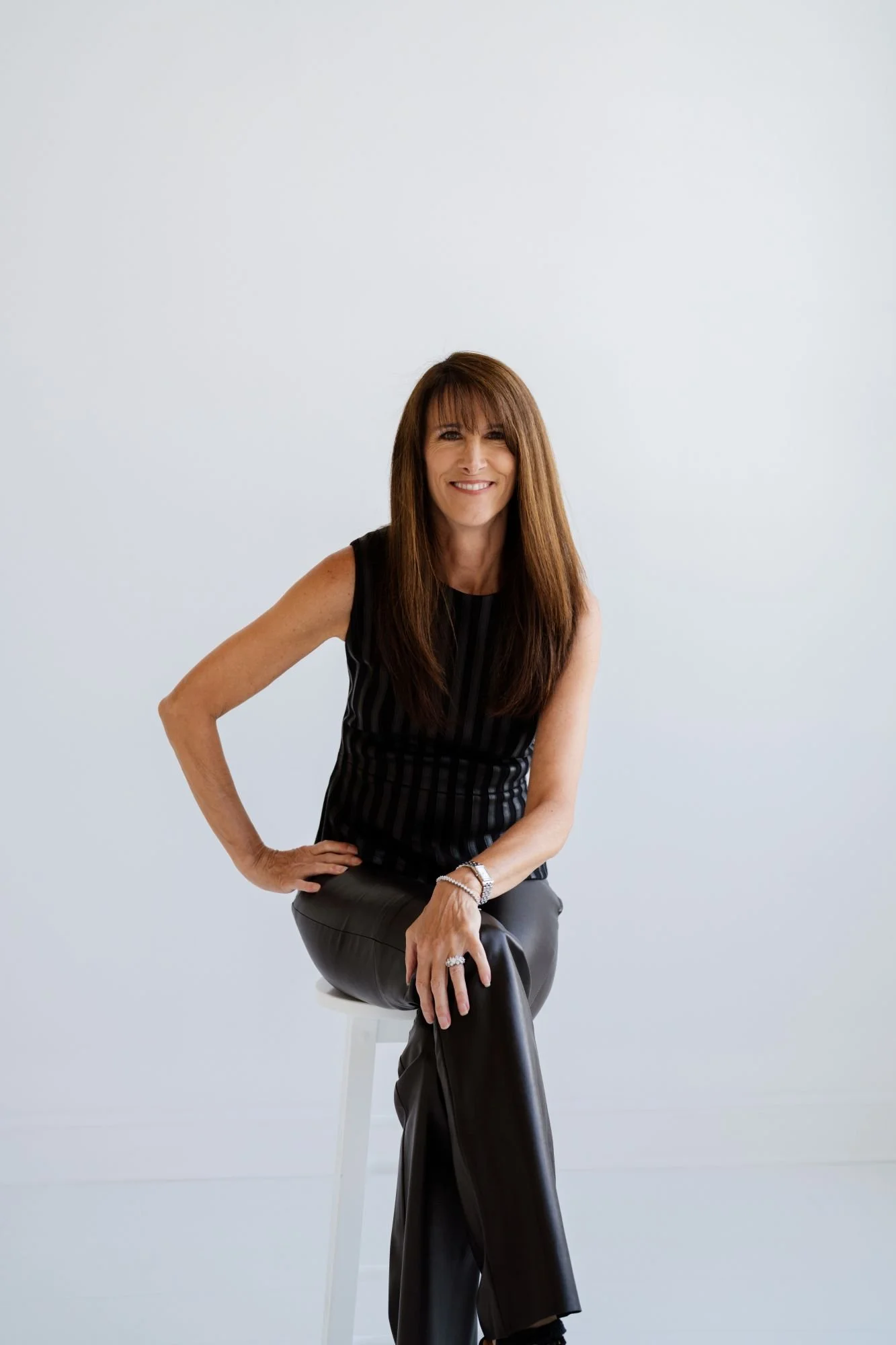A woman with long, brown hair, wearing a sleeveless striped black top and black leather pants, smiling while sitting on a white stool against a plain white background.