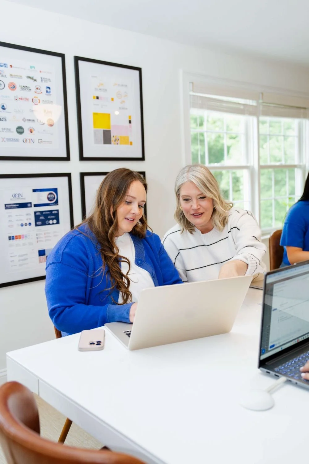 Two women working together at a white desk in a bright office, looking at a laptop screen with framed artwork on the wall behind them and windows letting in natural light.