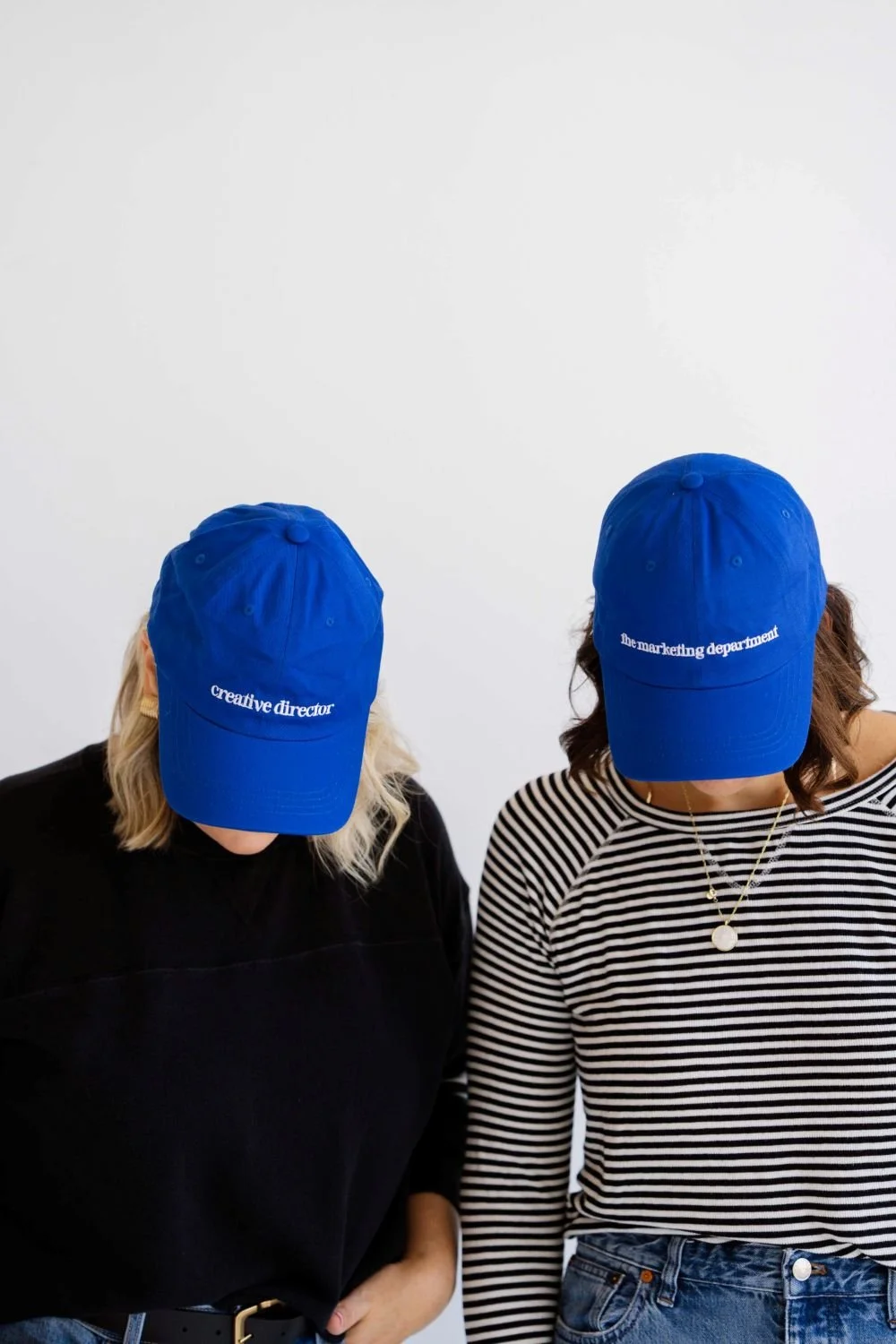 Two women wearing blue caps with white text, one says 'creative director' and the other 'the marketing department', both are looking down, against a plain white background.