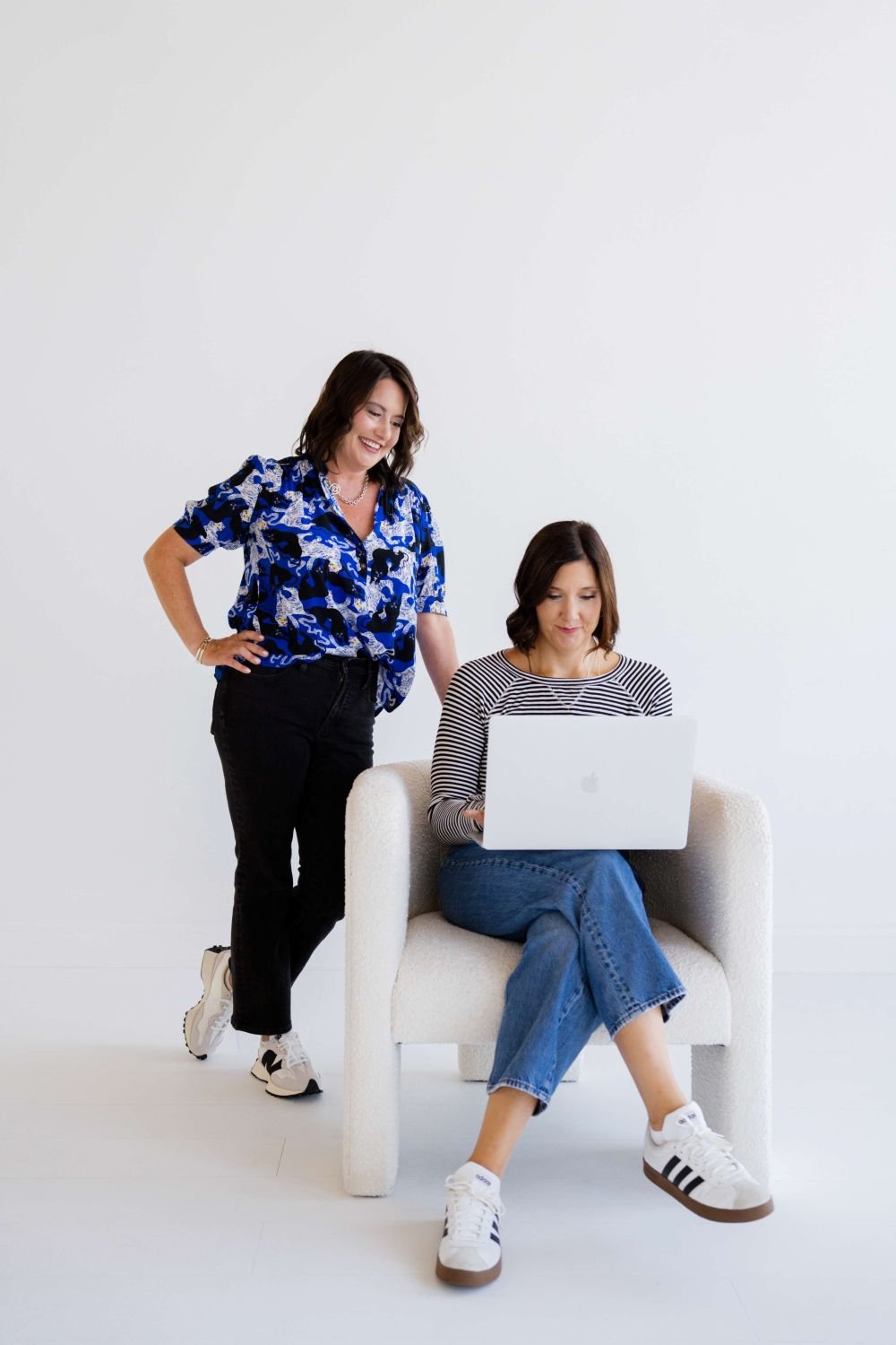 Two women with dark brown hair, one sitting on a cream armchair using a laptop and the other standing beside her, smiling. The standing woman wears a blue patterned blouse and black pants, while the seated woman wears a striped long-sleeve top and blue jeans, with white sneakers.