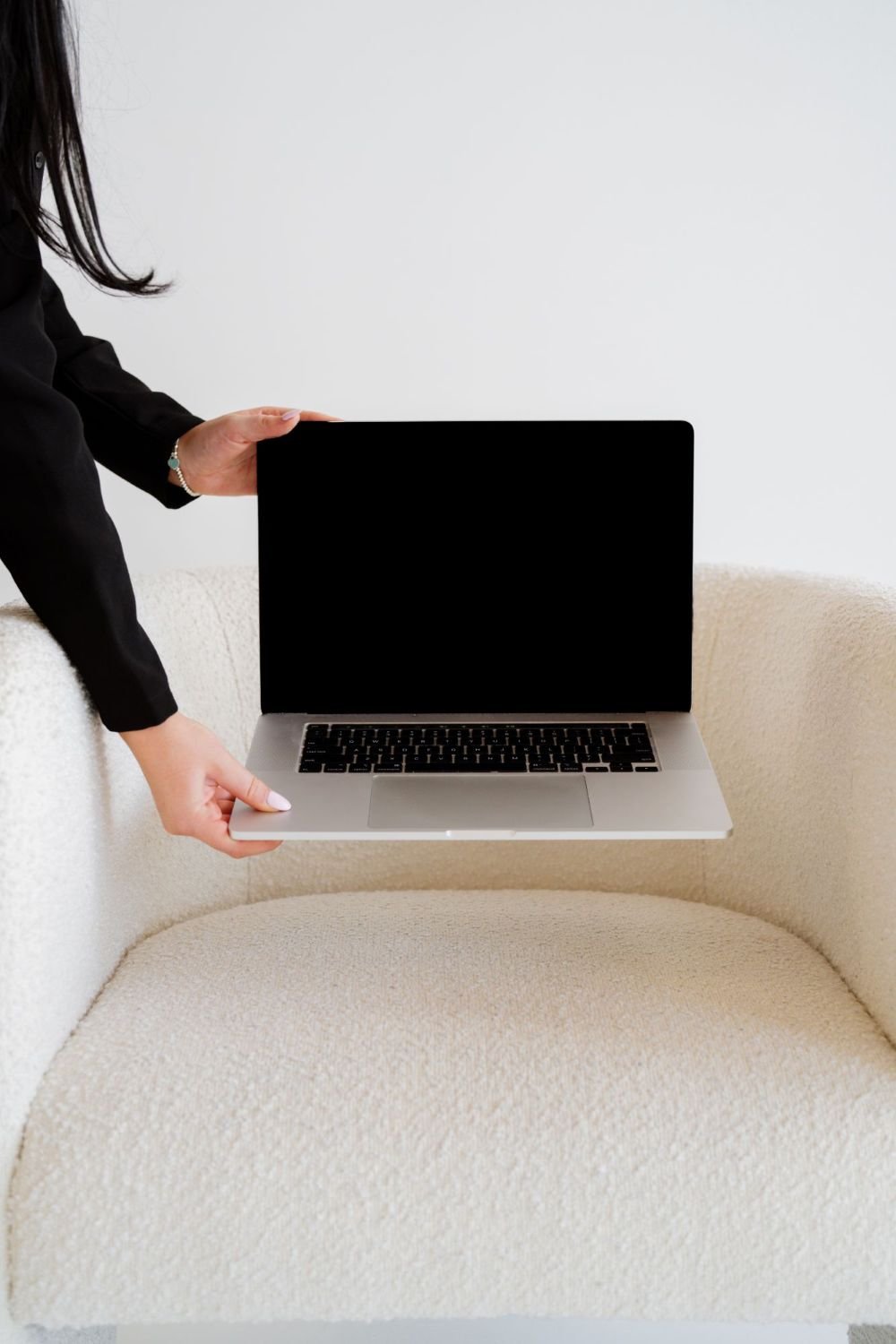 Person holding a silver laptop with a black screen, sitting on a beige sofa with a white wall background.