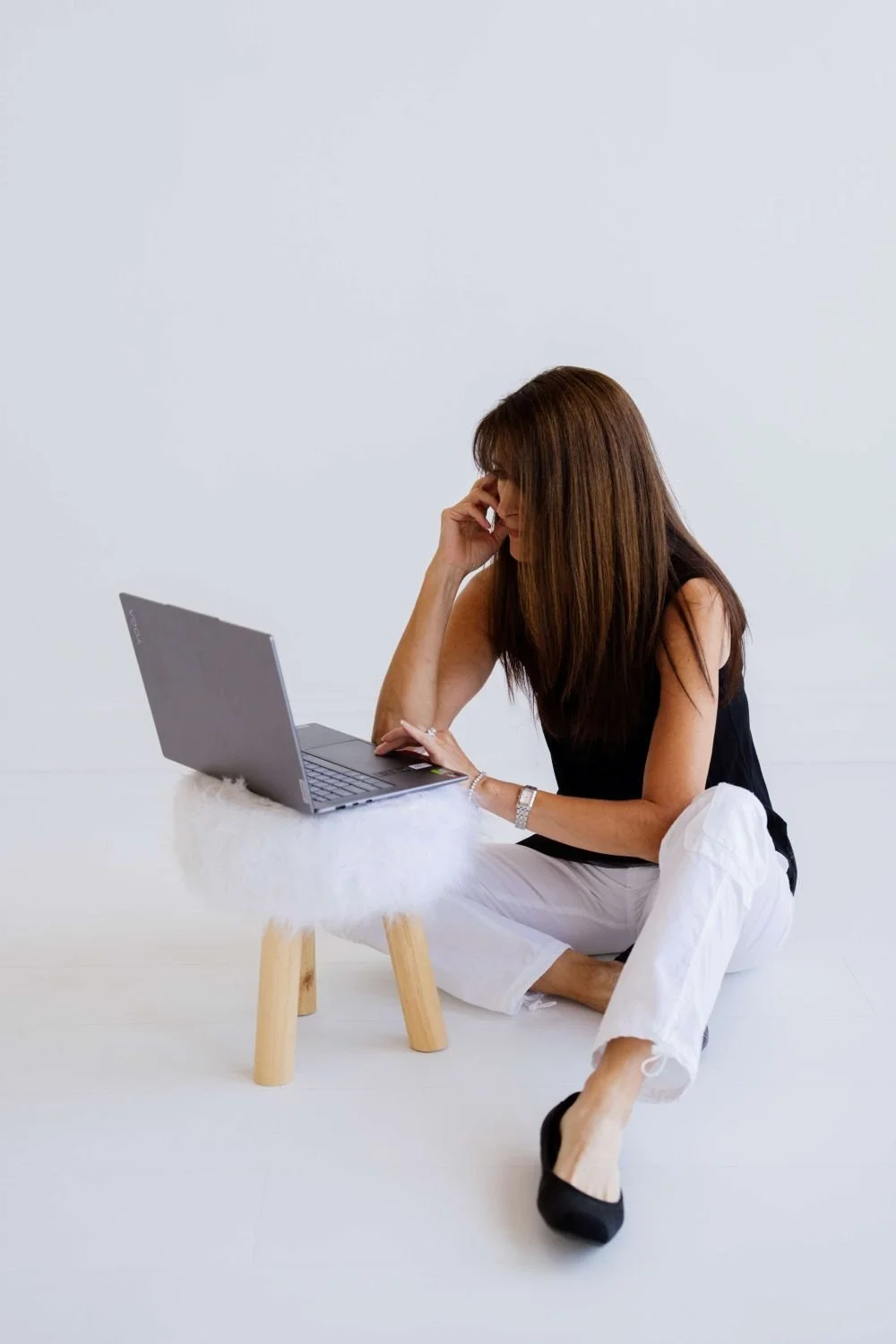 Woman sitting on the floor, looking at a laptop, with one hand on her forehead, in a minimal white room.