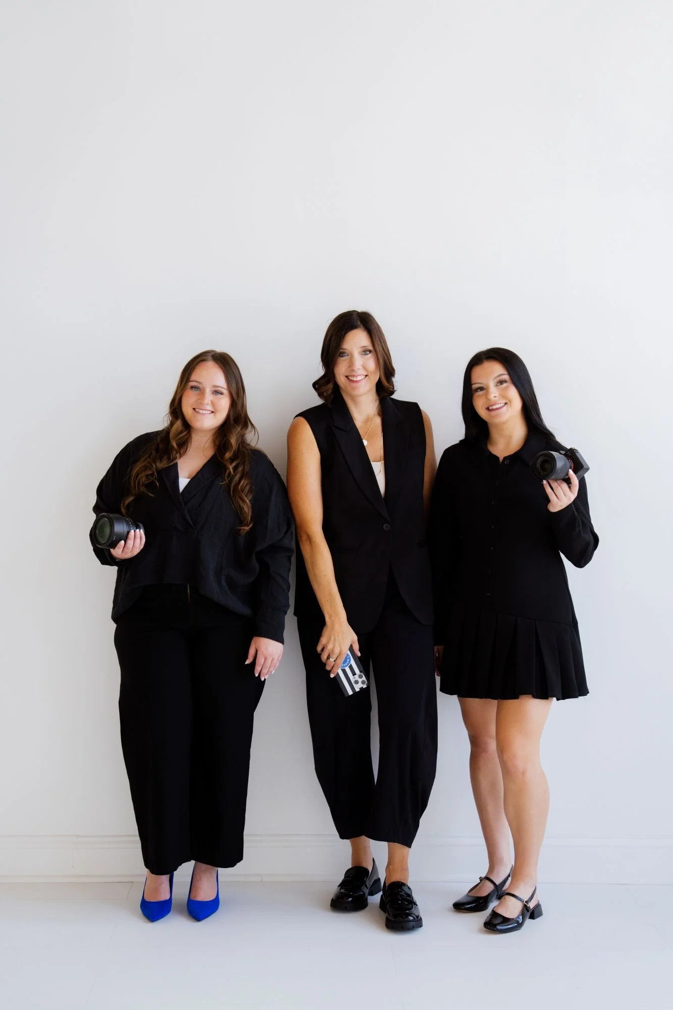 Three women standing against a plain white wall, smiling. Two women on the left and right are holding cameras, and the woman in the middle is wearing a sleeveless blazer and holding a motion picture clacker.