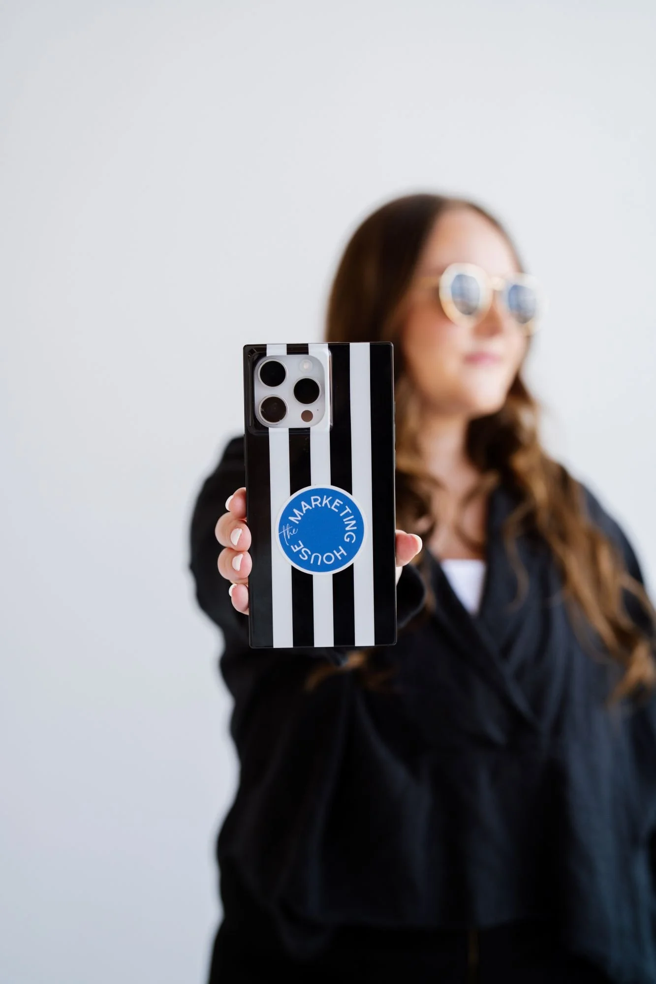 A woman taking a selfie with her smartphone, which has a black and white striped case with a circular blue sticker that says 'The House of the Marketing.' The woman is wearing sunglasses and a black jacket, and has long brown hair. The background is a plain white wall.