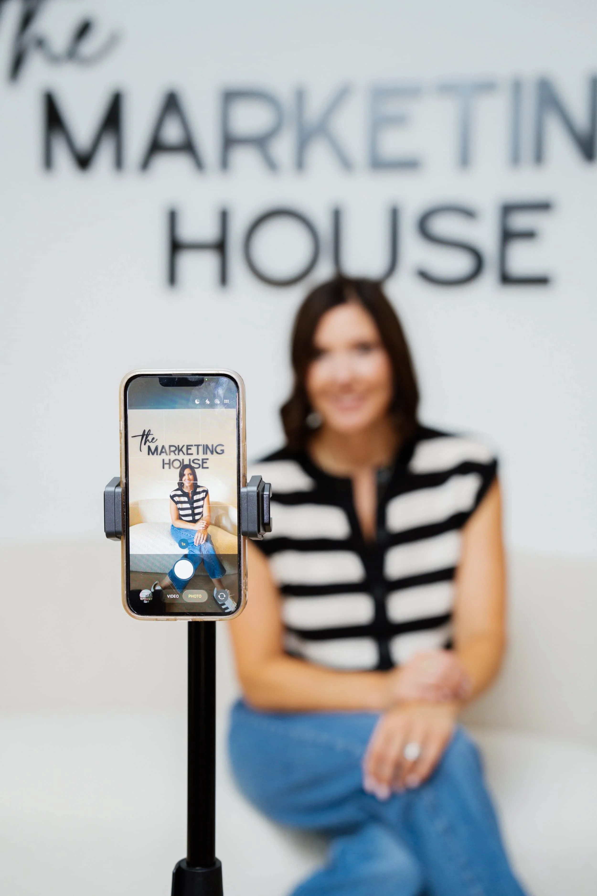 A woman is sitting against a white wall with the words 'the MARKETING HOUSE' written on it. She is wearing a black and white striped shirt and jeans, and is smiling with her arms crossed. In front of her, a smartphone mounted on a stand is taking a photo of her, displaying her on the screen.