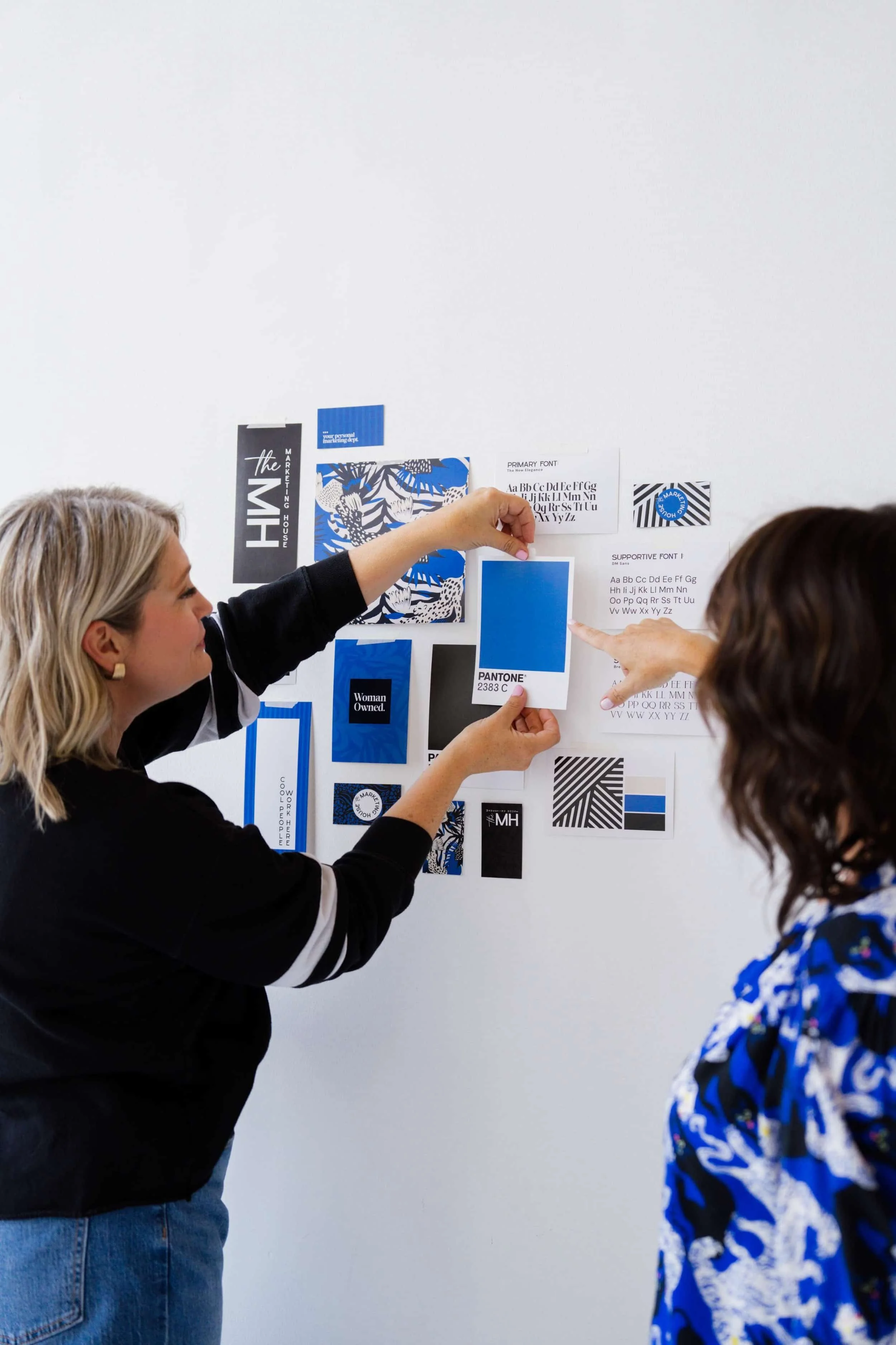 Two women discussing a color palette and font selection on a wall with various printed design samples.