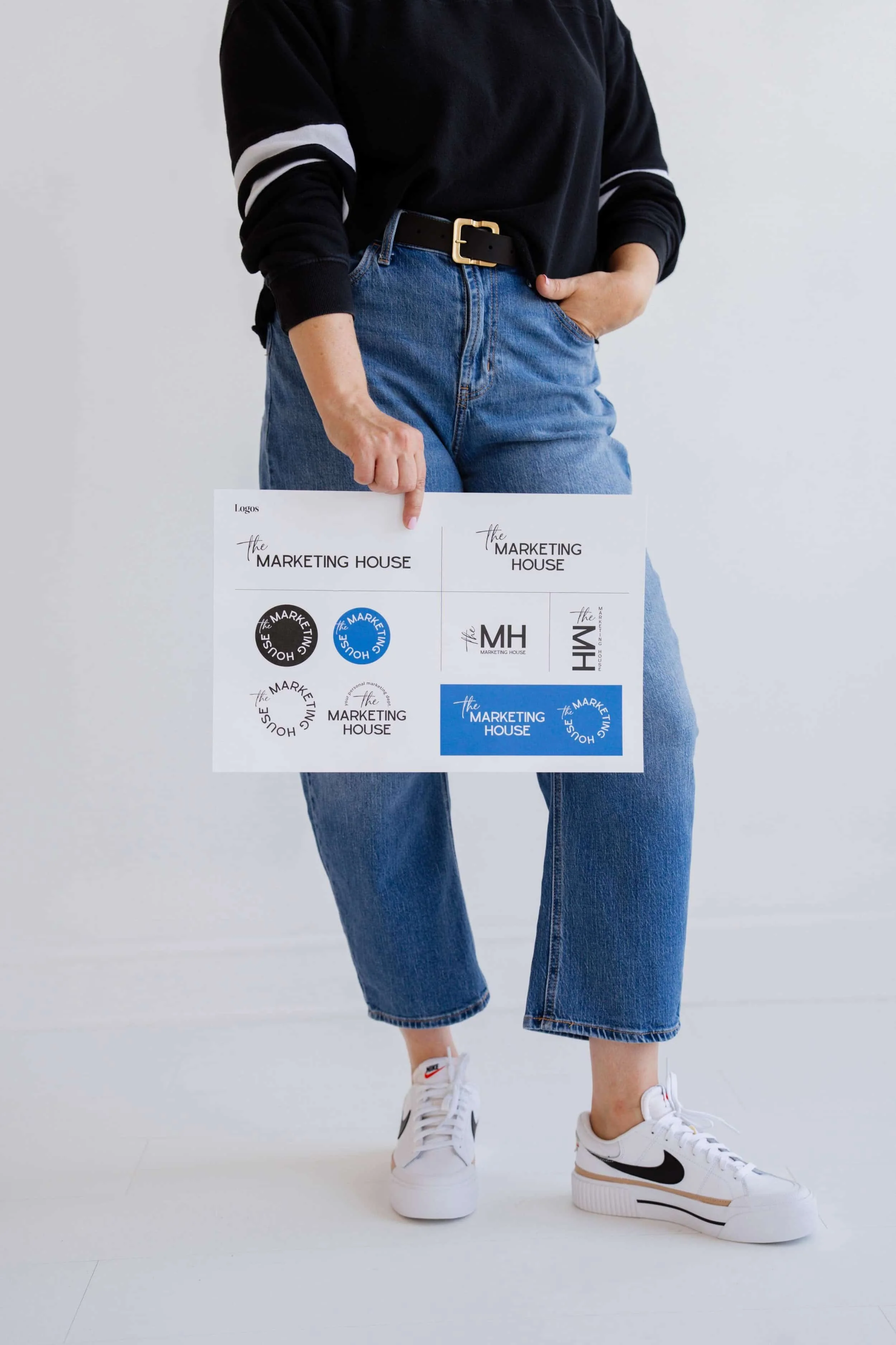 Person wearing a black long-sleeve shirt with white stripe on sleeves, blue jeans, and white sneakers with black Nike swoosh, standing against a plain light gray background, holding a sheet of branding logos.