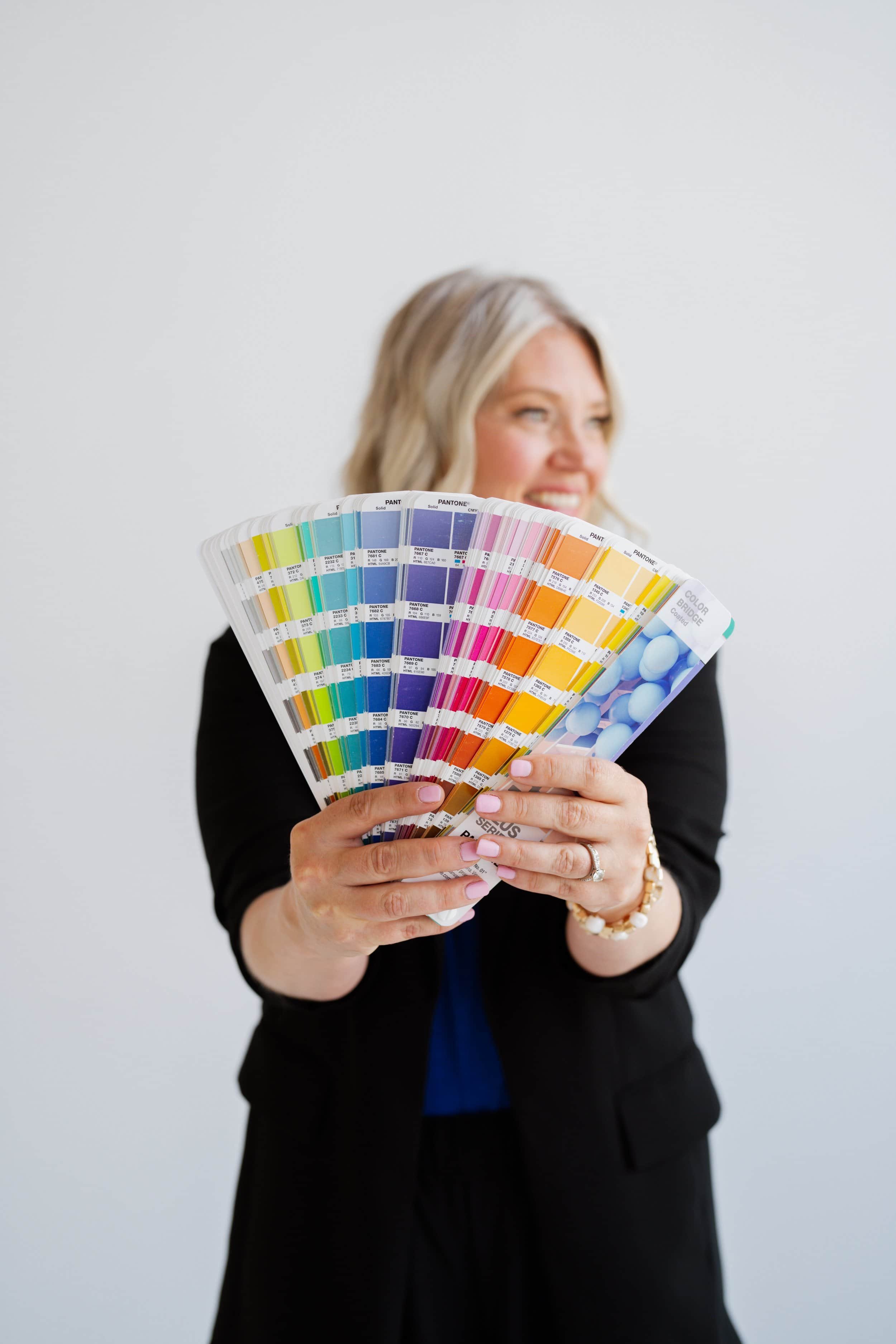 A woman holding a color sample fan with various shades of blue, purple, pink, orange, and yellow, smiling and wearing a black jacket with a gold watch and bracelet, against a plain white wall.