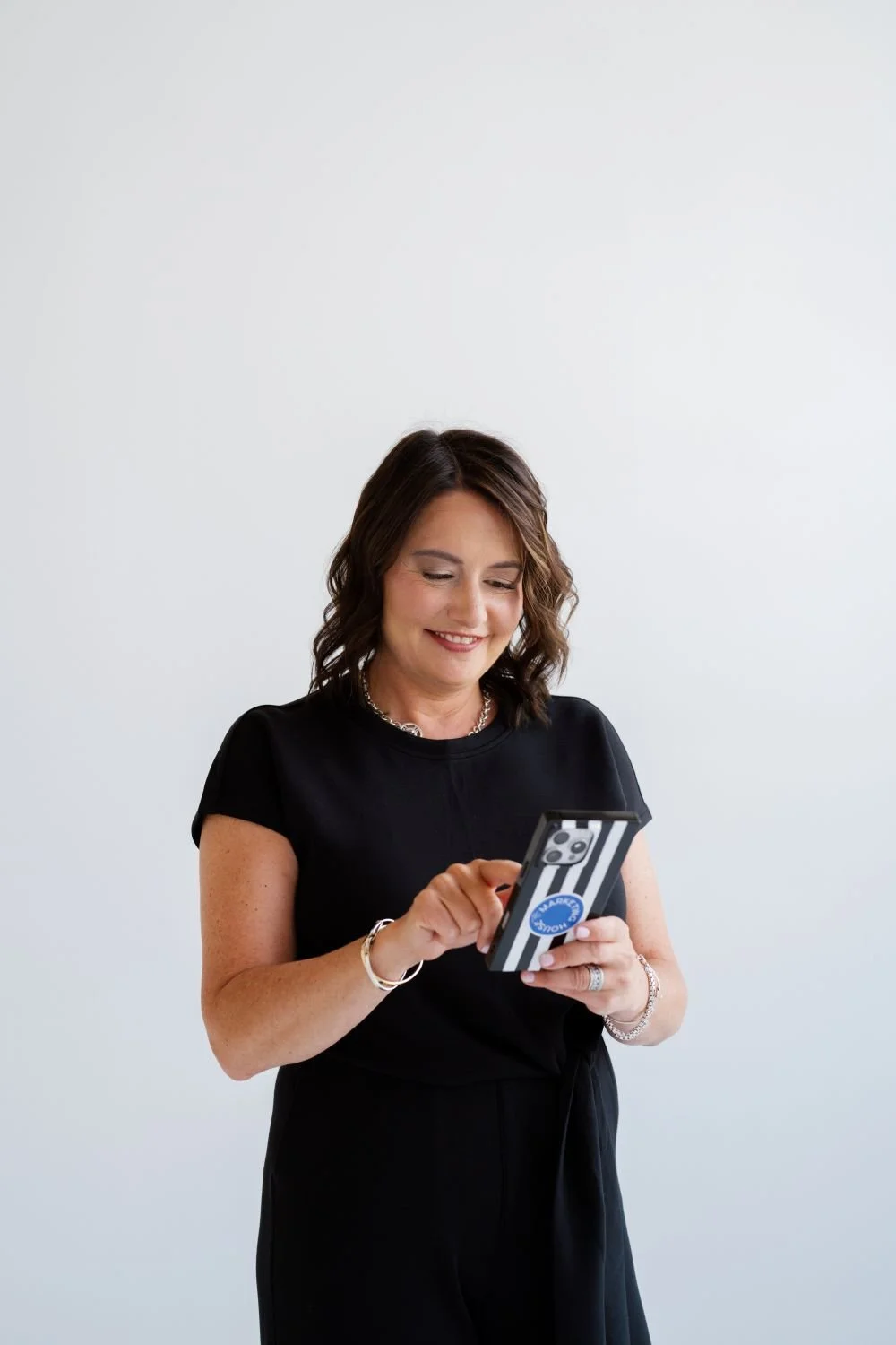 A woman with shoulder-length brown hair wearing a black top and jewelry, smiling while looking at her smartphone with a black and white striped case and a sticker that says "you rock."