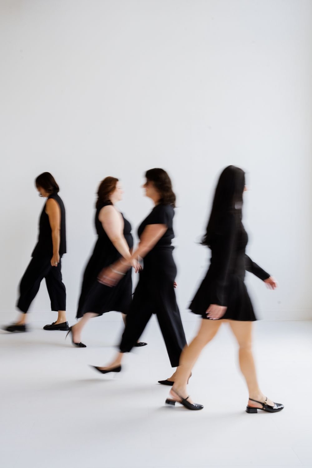 Four women in black clothing walking in a line on a white background.