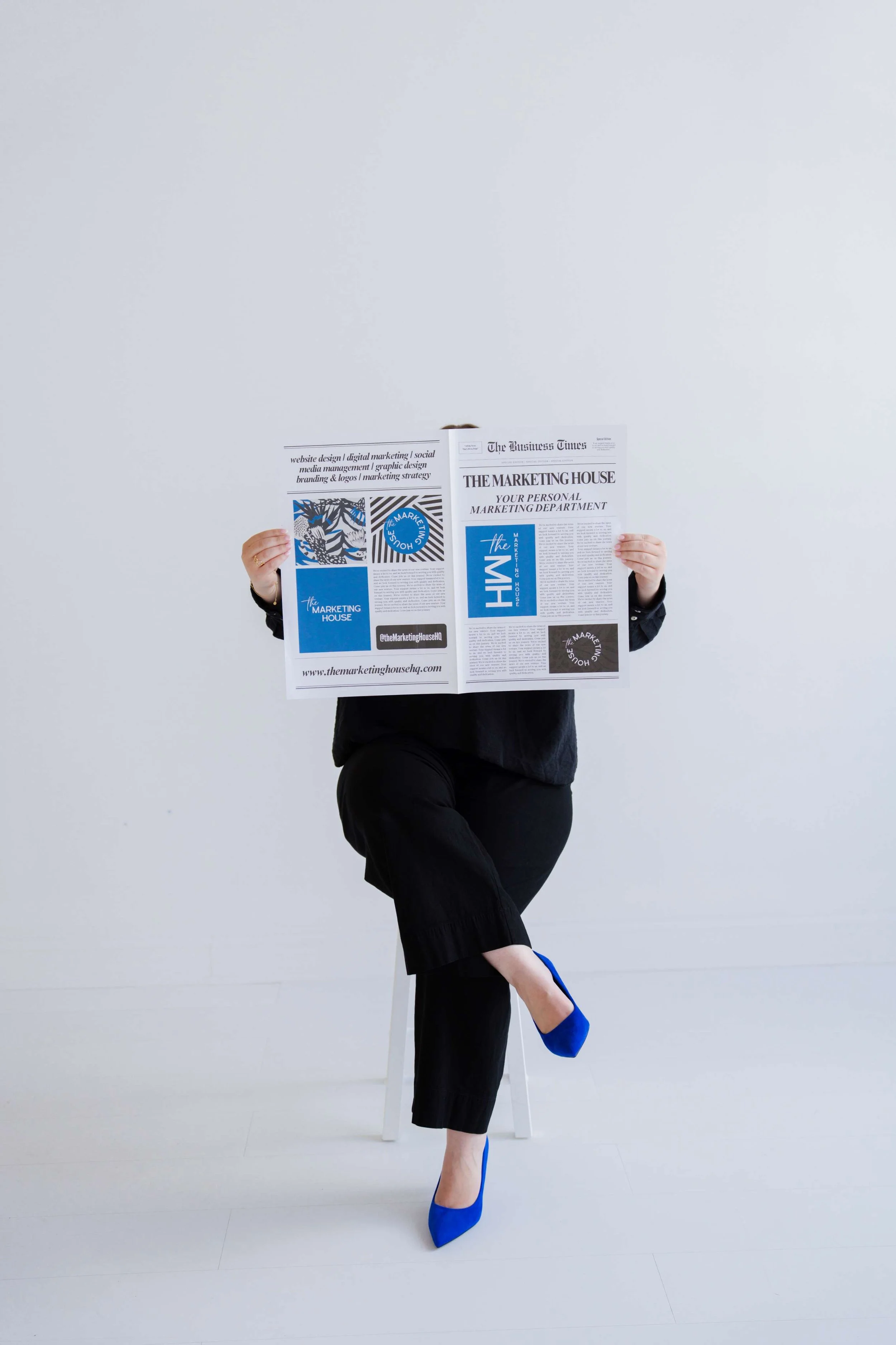 Person in black outfit and blue high heels sitting on a stool, holding a newspaper that covers their face against a plain white background.