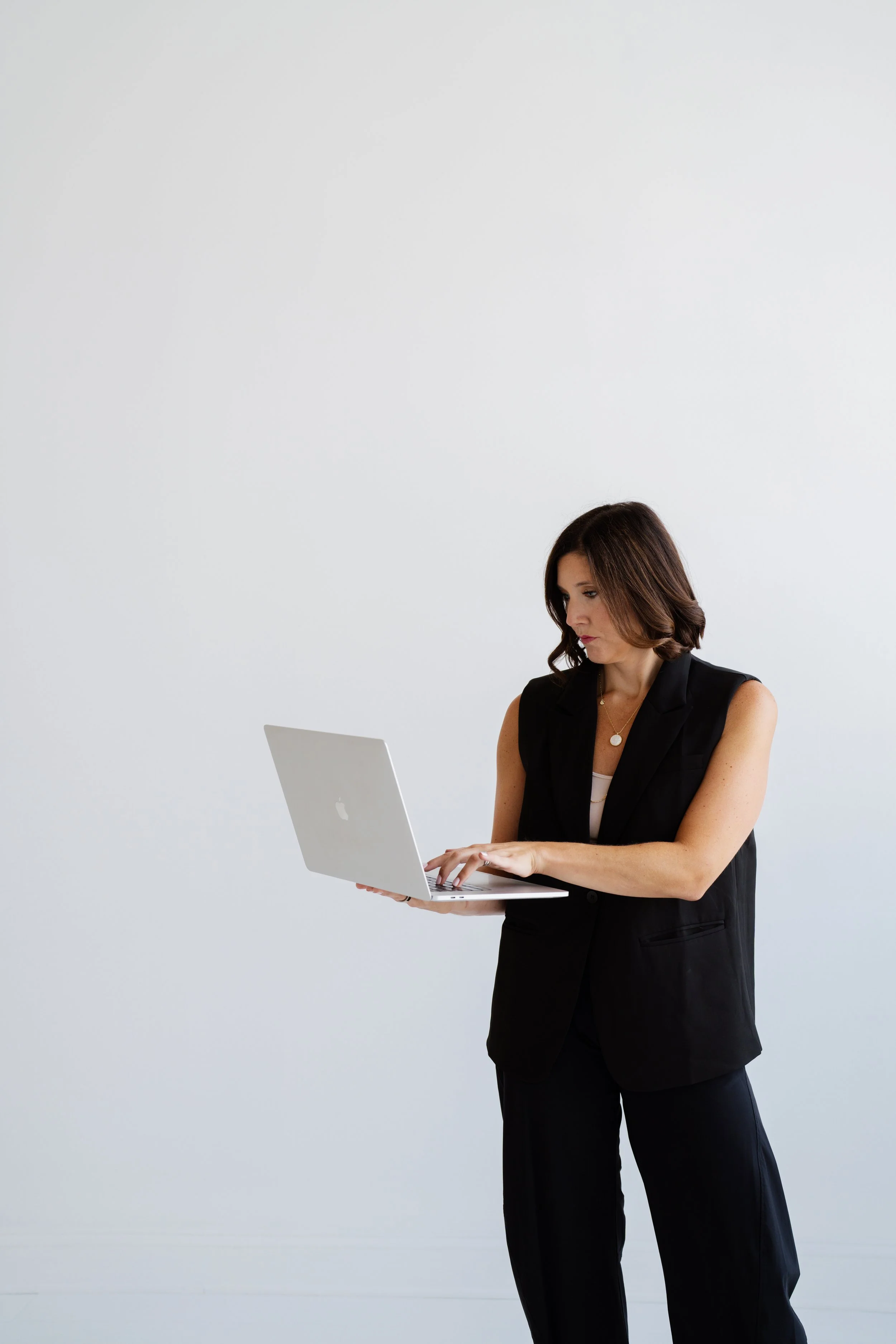 A woman with brown hair wearing a black sleeveless blazer and black pants, working on a silver laptop against a plain light gray background.