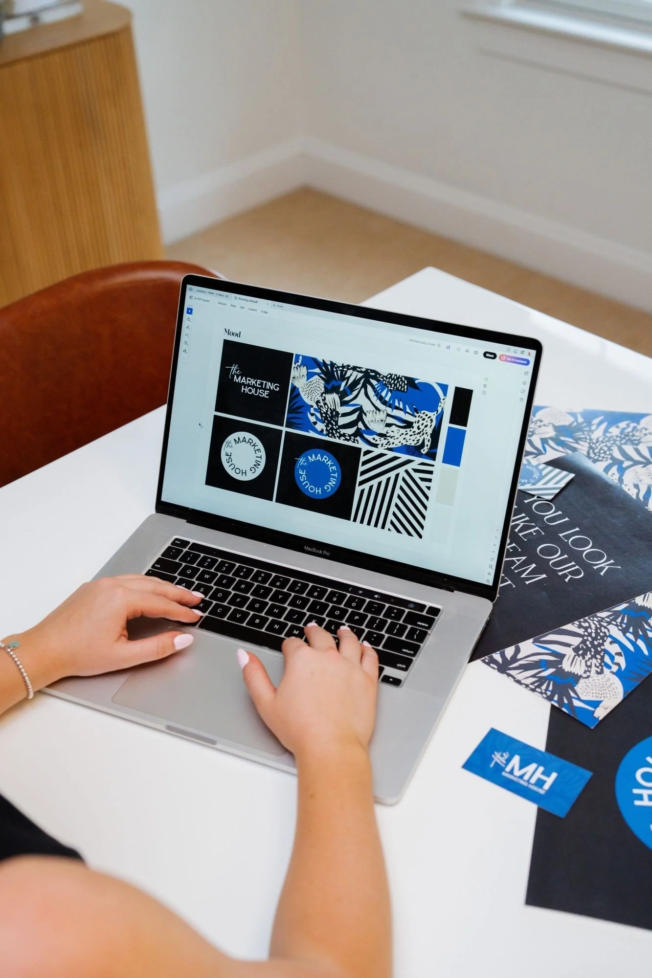 A person working on a laptop displaying graphic design layouts for a marketing project, with branded materials and brochures on a white table.
