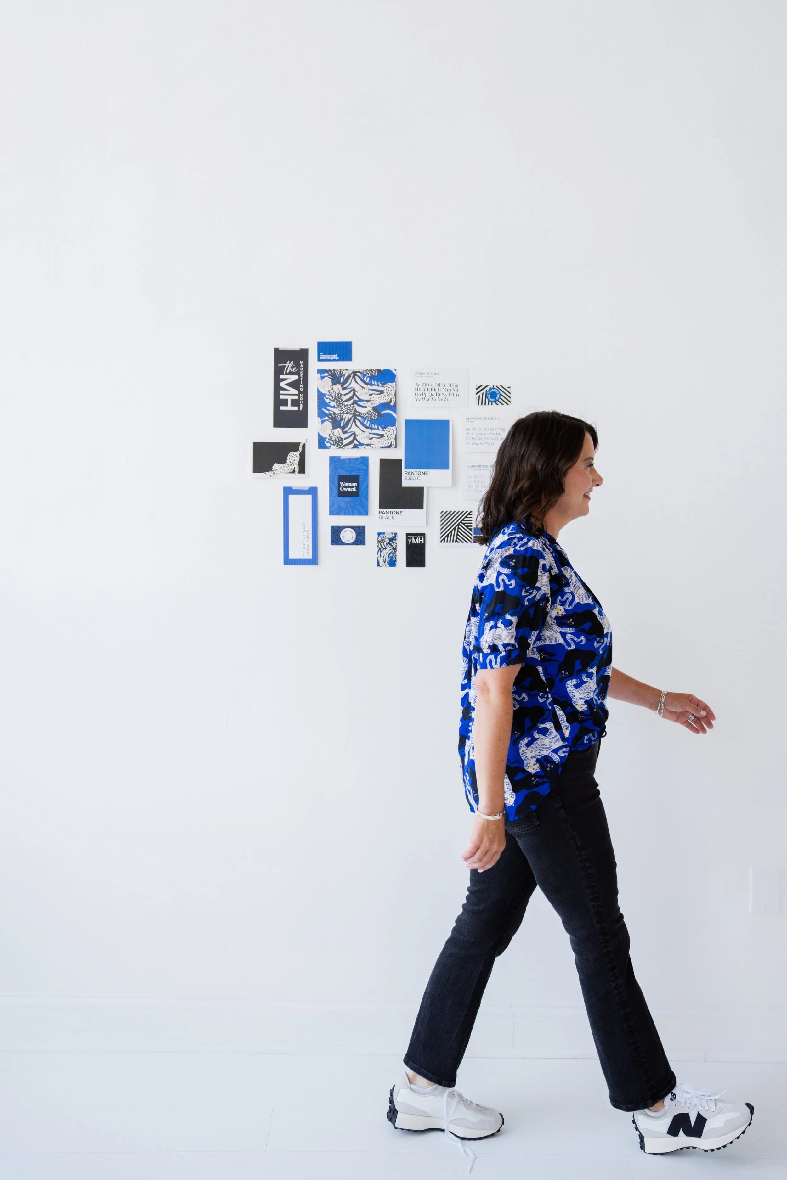 A woman walking in a white room with a wall of blue and black graphic posters in the background. She has shoulder-length dark hair, is smiling, and wearing a blue and white patterned shirt, black jeans, and white sneakers.