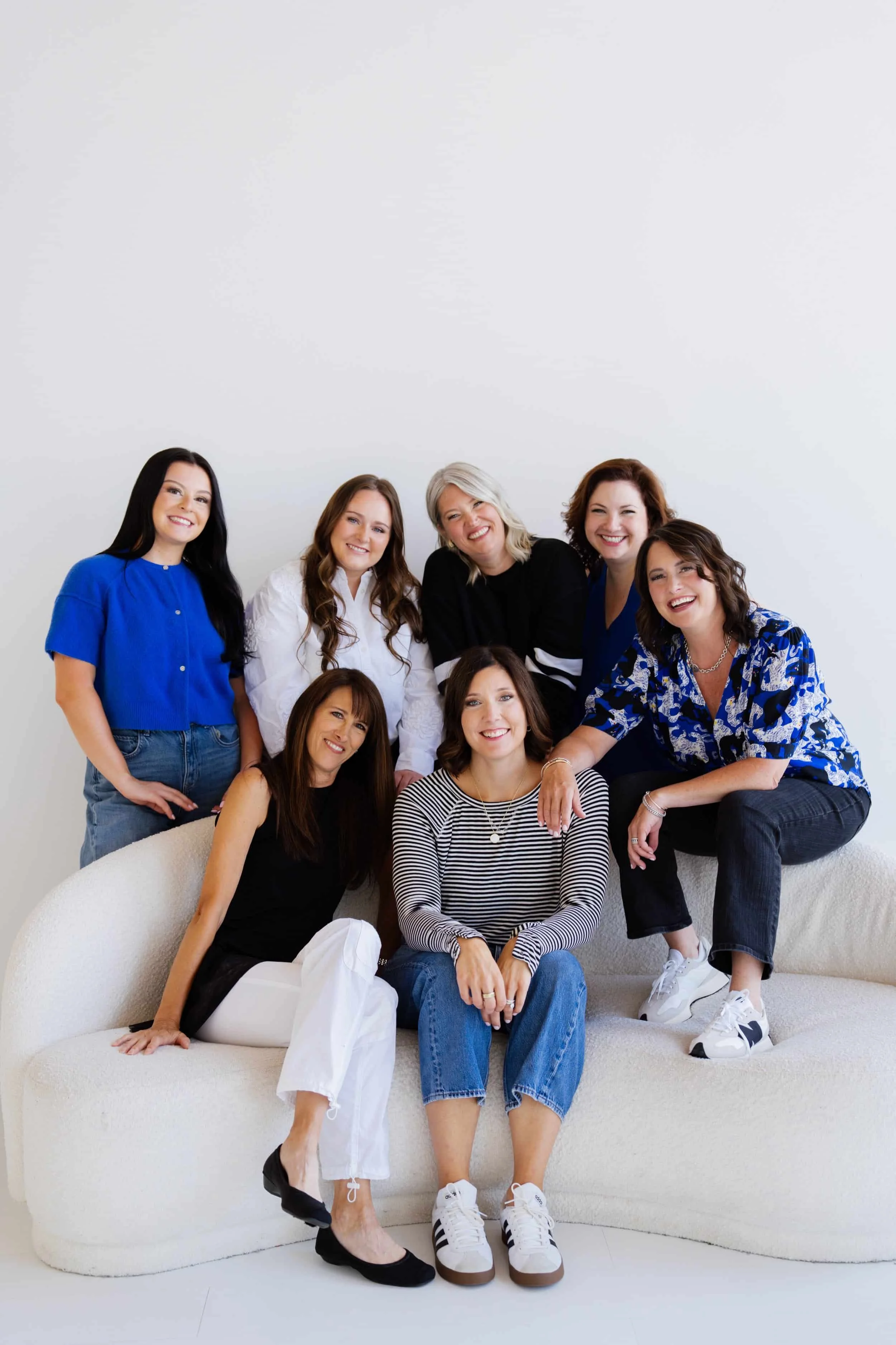 Group of seven women smiling together in a minimalistic, white room, posing on and around a white curved sofa, dressed casually in colorful tops and jeans.