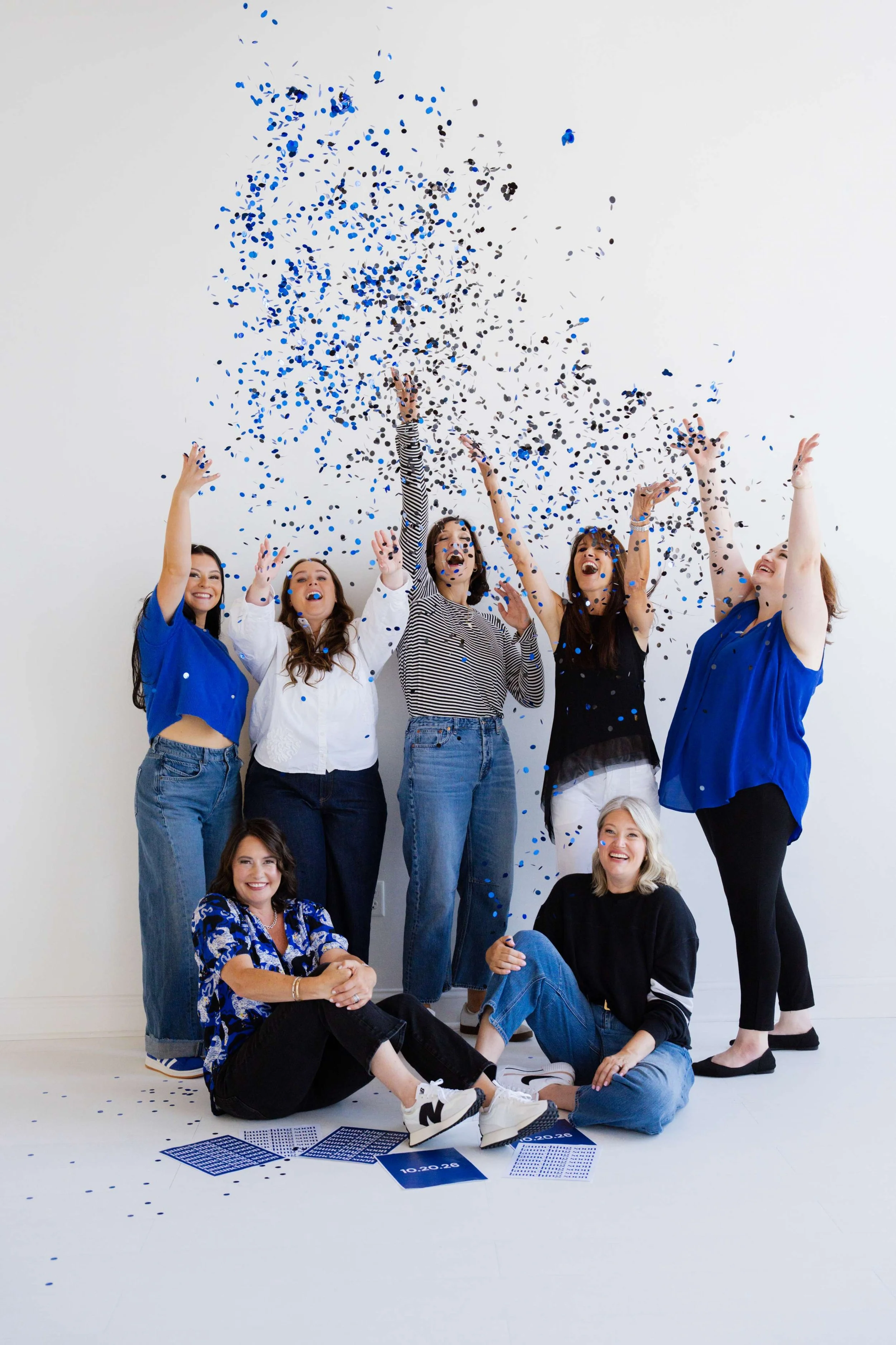 Group of smiling women celebrating with blue and black confetti in a photography studio with white walls.
