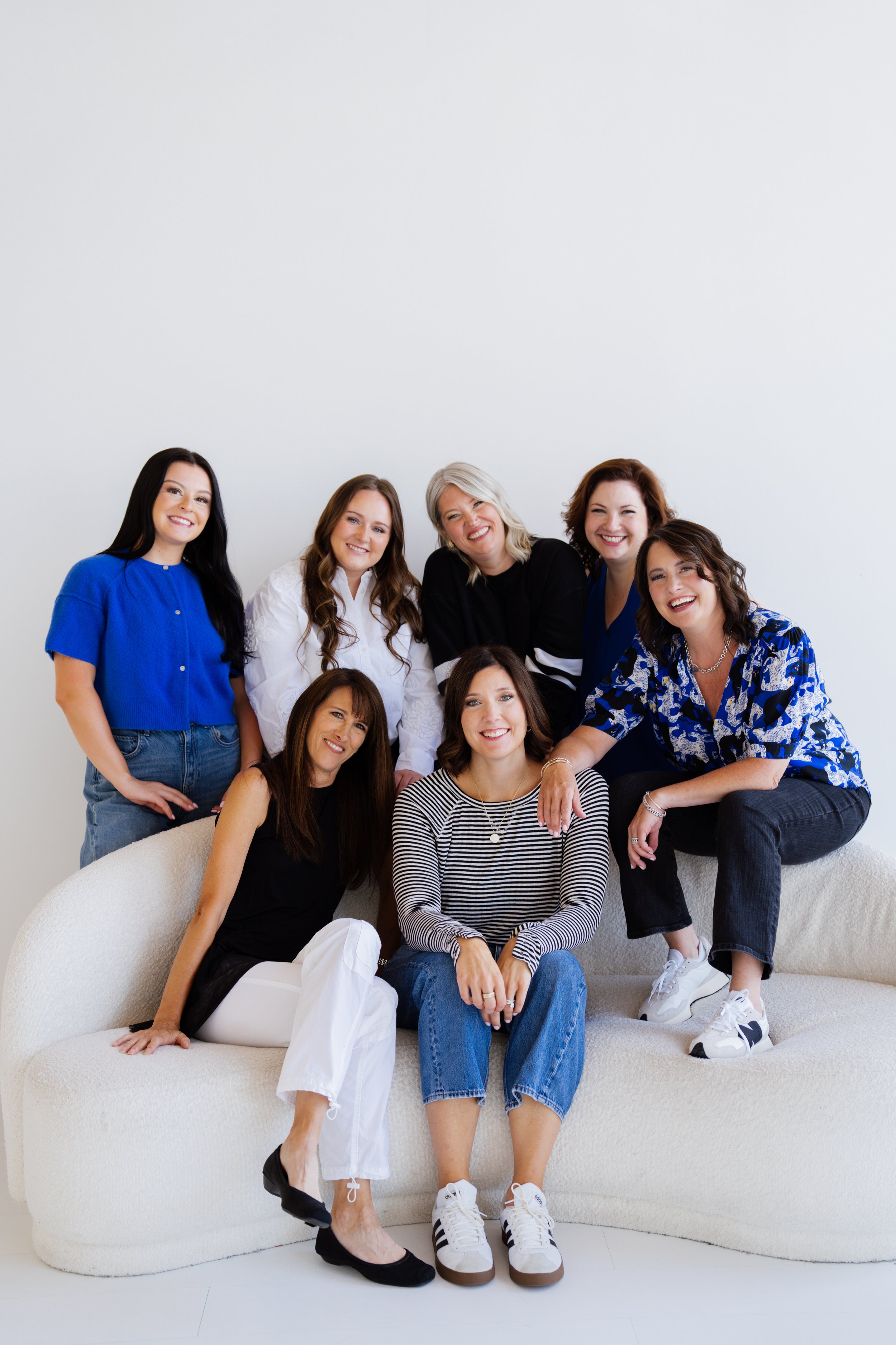 Group of nine women smiling and posing together on a white couch against a plain white wall.