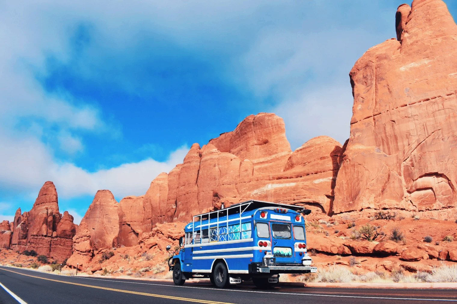 A blue RV parked on the side of a paved road with red rock formations and a partly cloudy blue sky in the background.