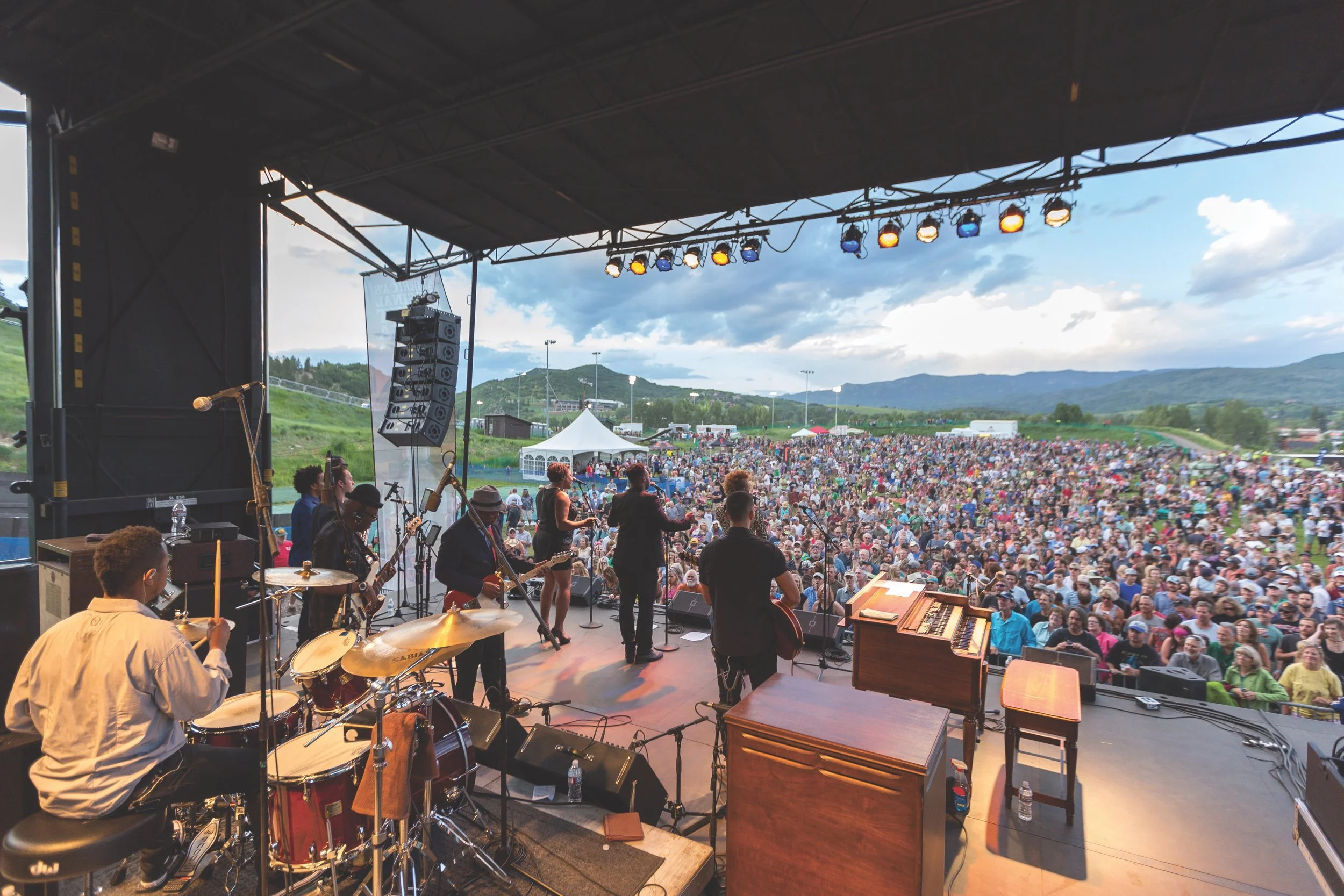 A live outdoor musical performance with a band on stage and large audience in a scenic mountain landscape.