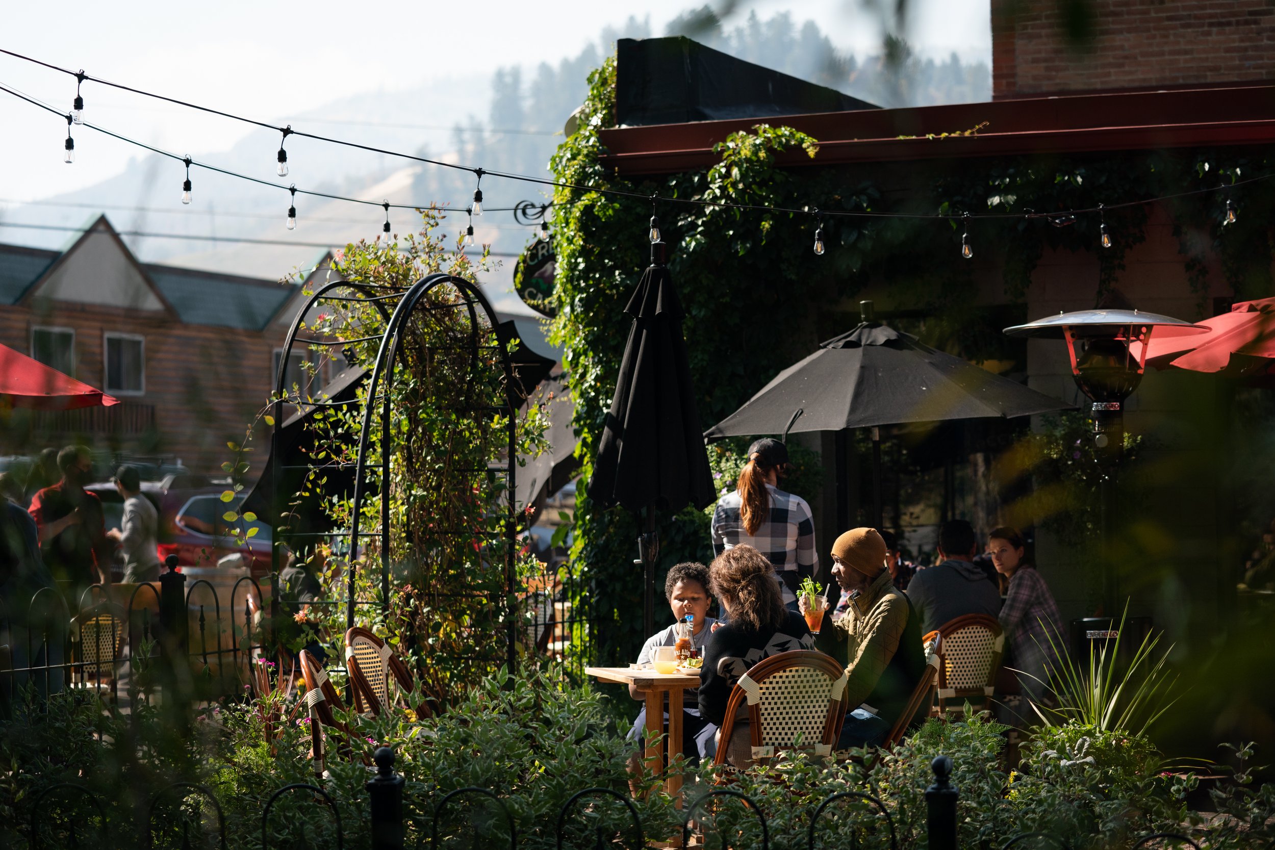 People dining outdoors at a restaurant, shaded by umbrellas, with string lights hanging overhead and a mountain in the background.