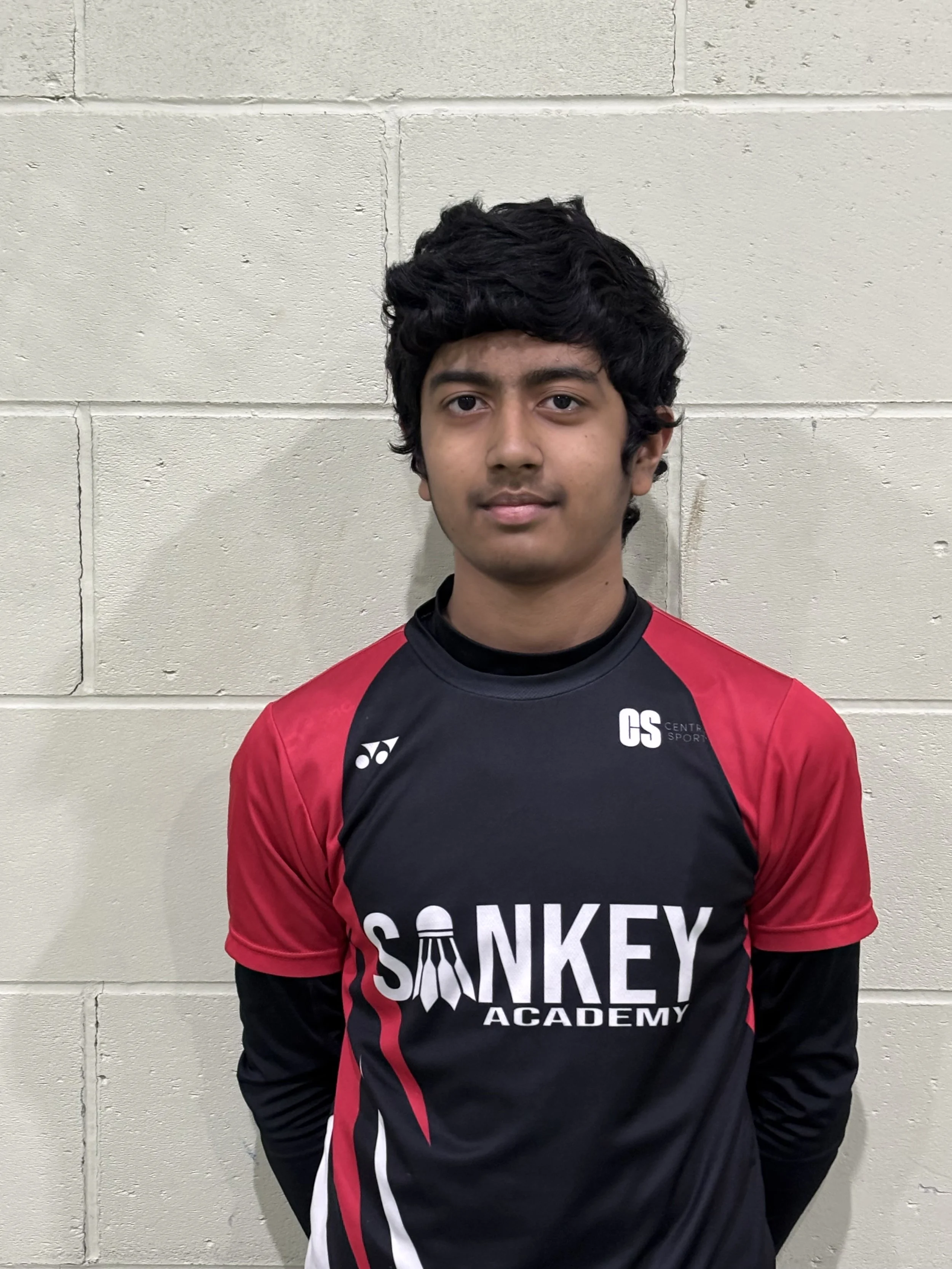 A young boy wearing a black and red sports jersey standing against a plain white wall.