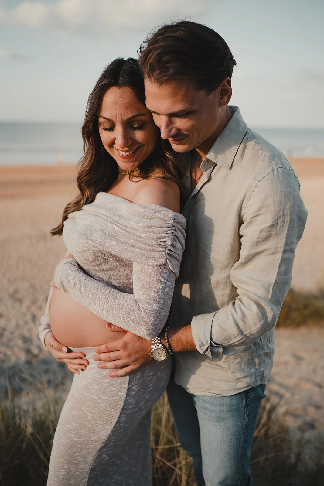 Un couple attend un bébé sur la plage, la femme est enceinte, ils se tiennent proches avec un sourire, la mer en arrière-plan.