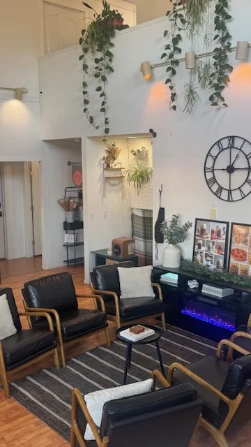 Cozy living room with black leather chairs, a black fireplace, and decorative plants hanging above. There's a large clock on the wall, framed pictures on a shelf, and a striped area rug.