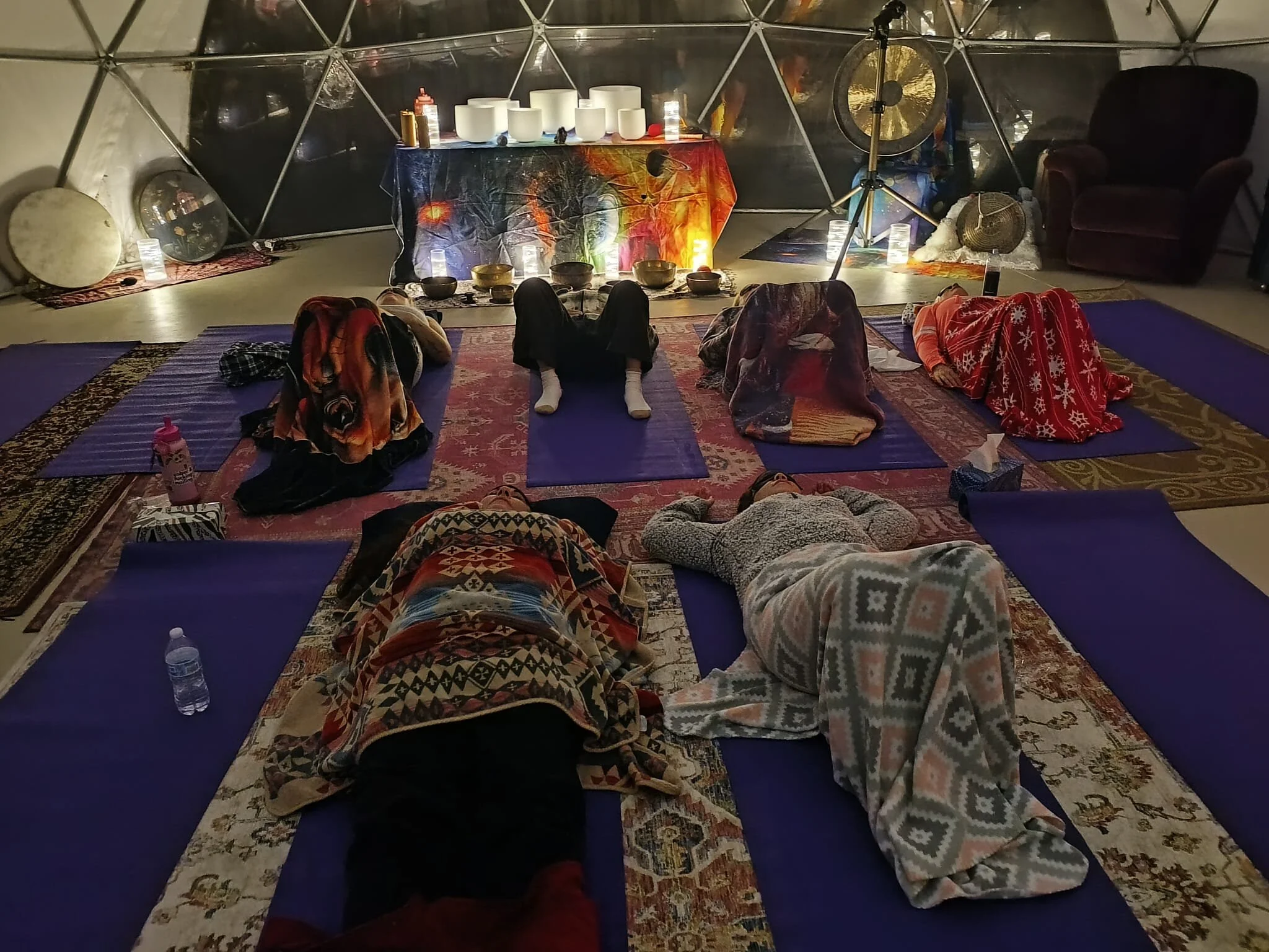 People lying on yoga mats in a dimly lit room with musical instruments and decorative items, seemingly participating in a relaxation or meditation session.
