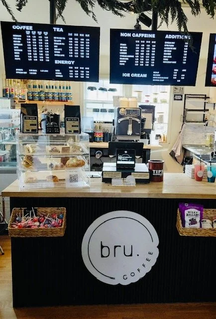 Coffee shop counter with digital menu boards displaying beverage options and prices, a coffee machine, baked goods in a display case, and cups on the counter. The logo 'bru. COFFEE' is on the front of the counter.