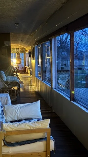 Interior of a sunroom or porch with a row of windows on the right, showing an outdoor deck and yard. There are chairs, a couch, and a lamp inside, with warm lighting creating a cozy atmosphere.