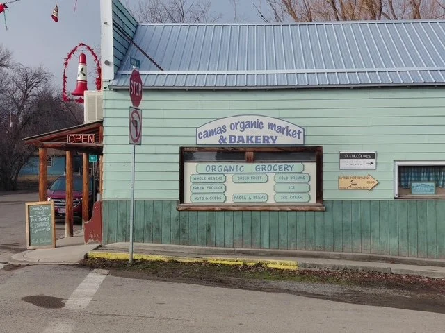 Exterior view of a small grocery store called 'Canvas Organic Market & Bakery' with a sign indicating 'Organic Grocery' and items like vegetables, pasta, and ice cream. A stop sign and an 'Open' sign are visible near the entrance.