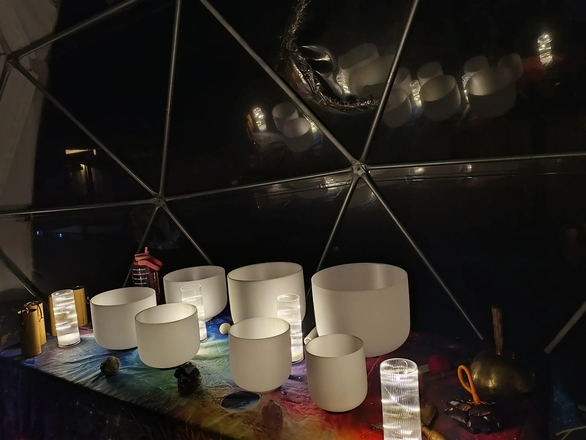 A display of musical instruments, including crystal singing bowls of various sizes, illuminated by small glowing lights, with a reflective black surface overhead and a colorful table covering.