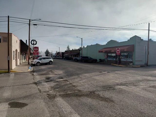 Empty street in a small town with parked cars, storefronts including a bakery, and holiday wreaths hanging on light poles, under an overcast sky.