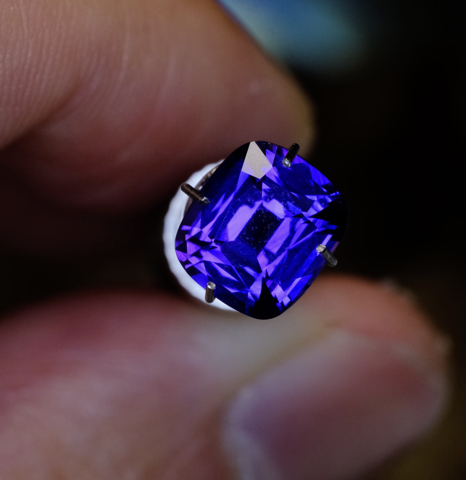 Macro view of a vivid blue, cushion-cut sapphire held between fingertips, secured by four prongs, with skin texture visible and a dark, softly blurred background.