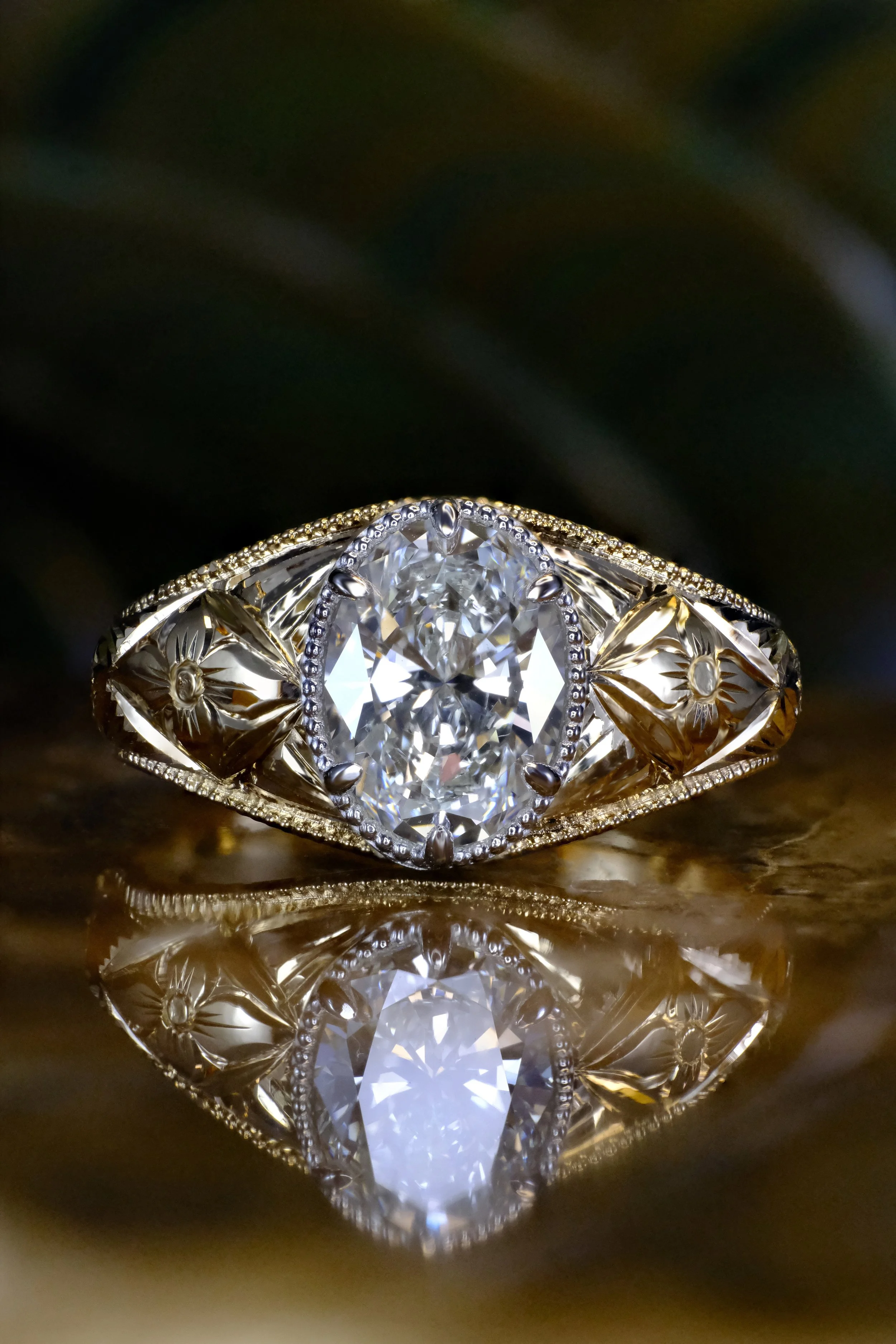 Yellow gold ring with oval diamond and engraved band, shown head-on with centered stone and reflection on a glossy surface.