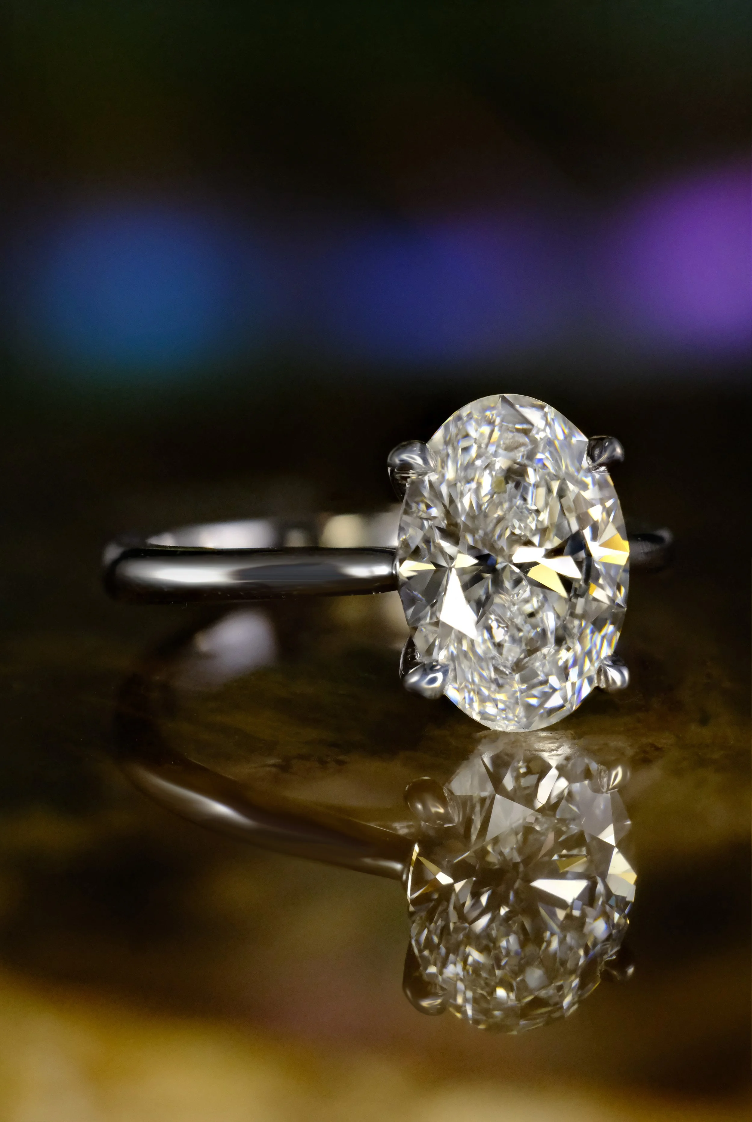 Side view of a white gold solitaire ring featuring an oval diamond held in a prong setting, shown with a smooth, polished band and a reflective surface beneath the ring.