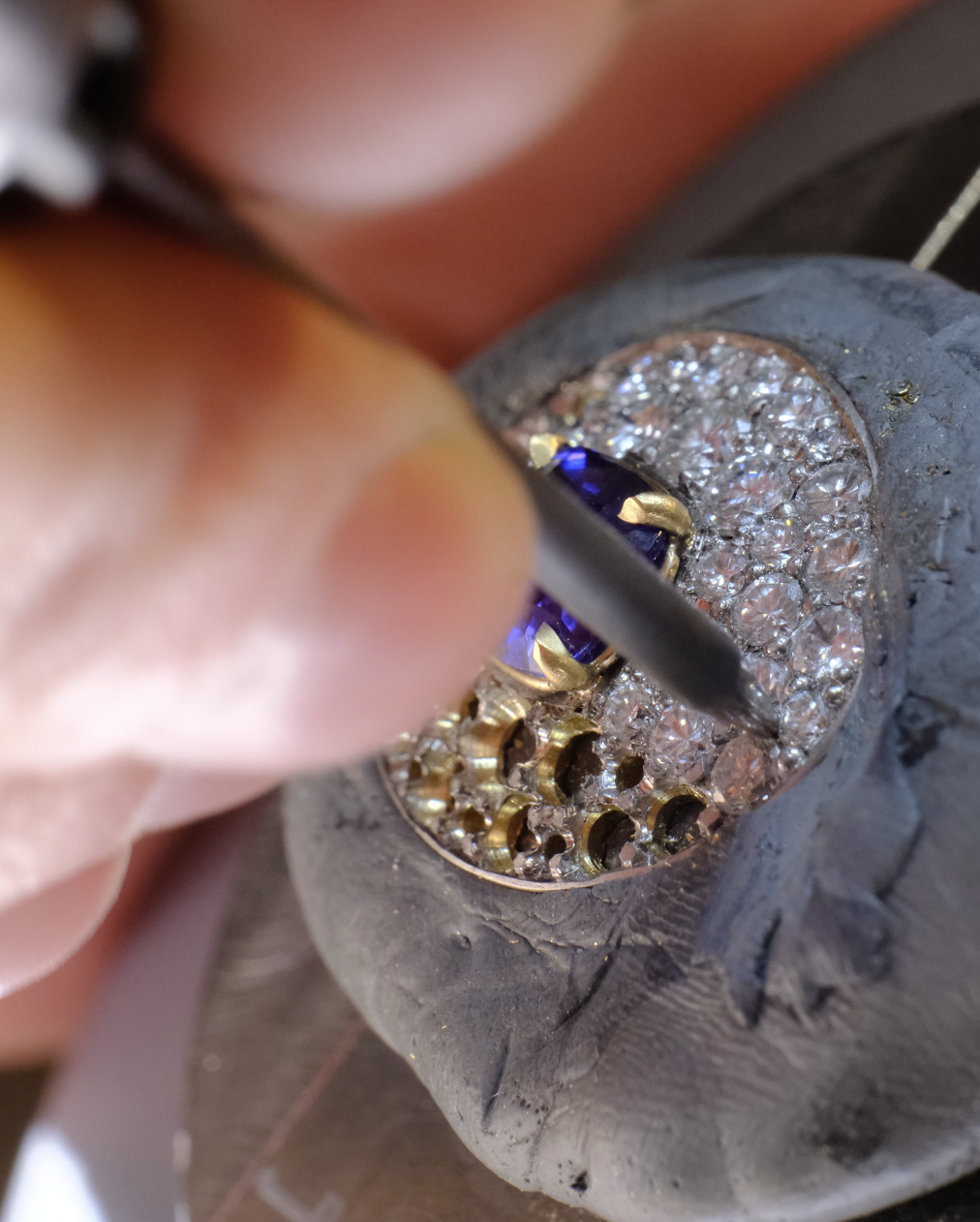 Close-up of a hand setting a blue sapphire in a gold mount, using a metal tool over pavé diamonds, with fingertips visible and gray setting compound surrounding the piece.
