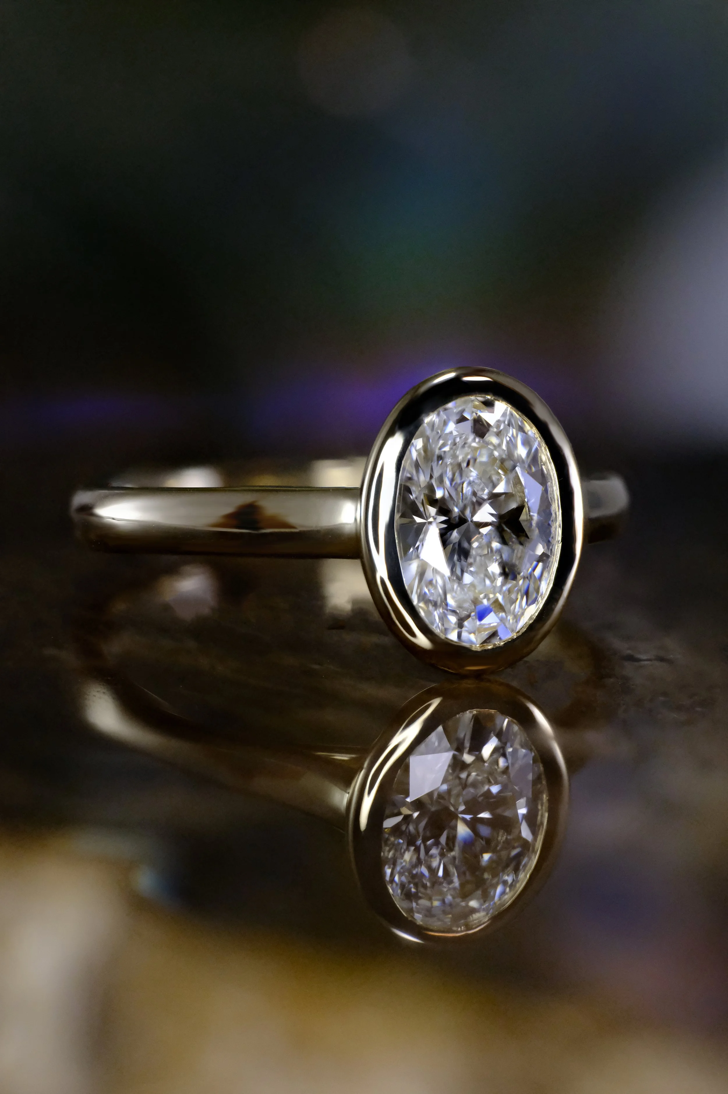 White metal (white gold or platinum) ring with an oval diamond in a smooth bezel setting, shown from the side with the band extending horizontally and the stone reflected on a glossy surface.