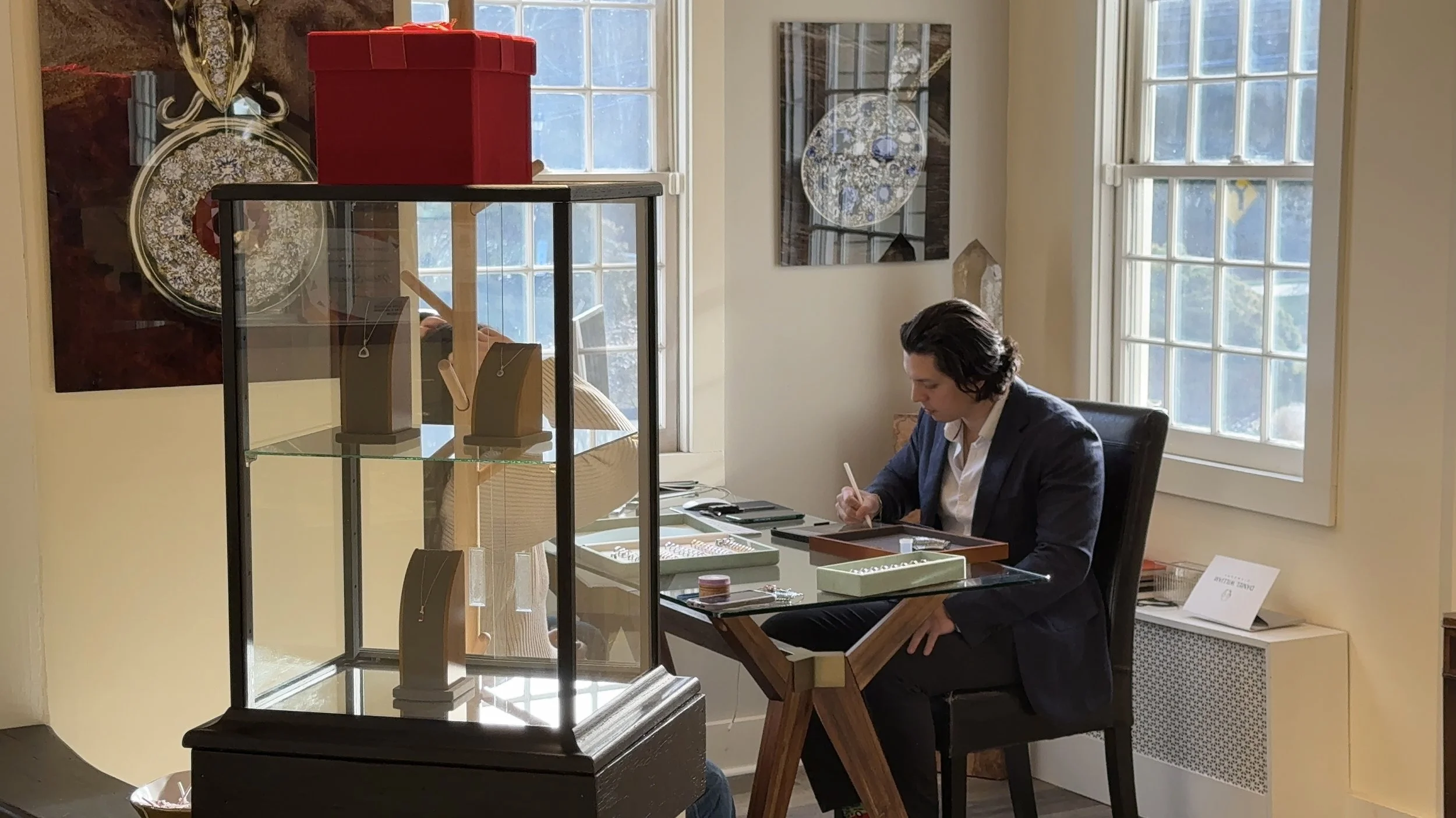 Jewelry designer seated at a glass desk sketching, surrounded by gemstone trays and display cases, with necklaces in a glass cabinet and daylight from large windows in a bright studio.