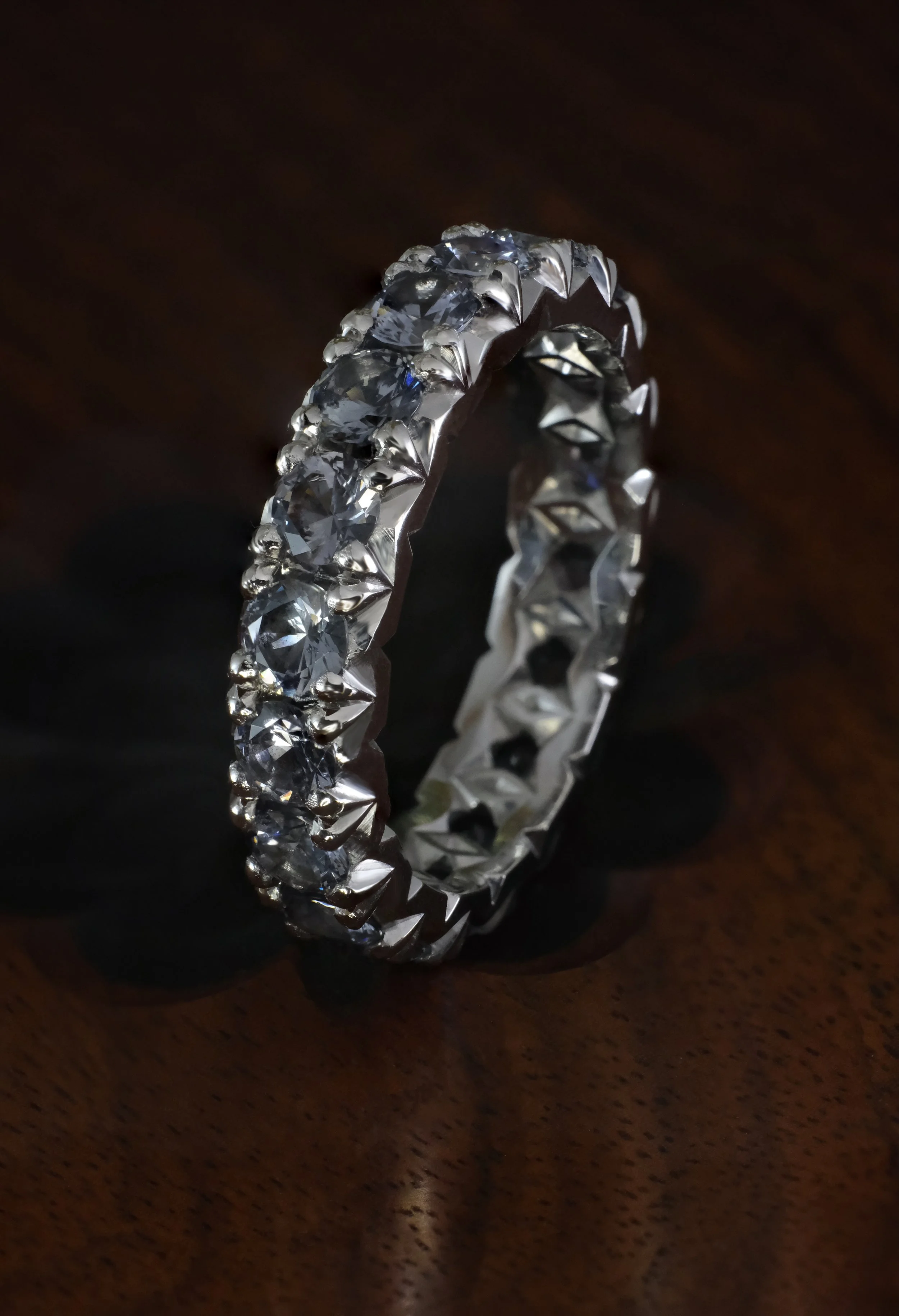 Platinum eternity band with round grey spinel stones, prong set all around, shown upright on a wooden surface.
