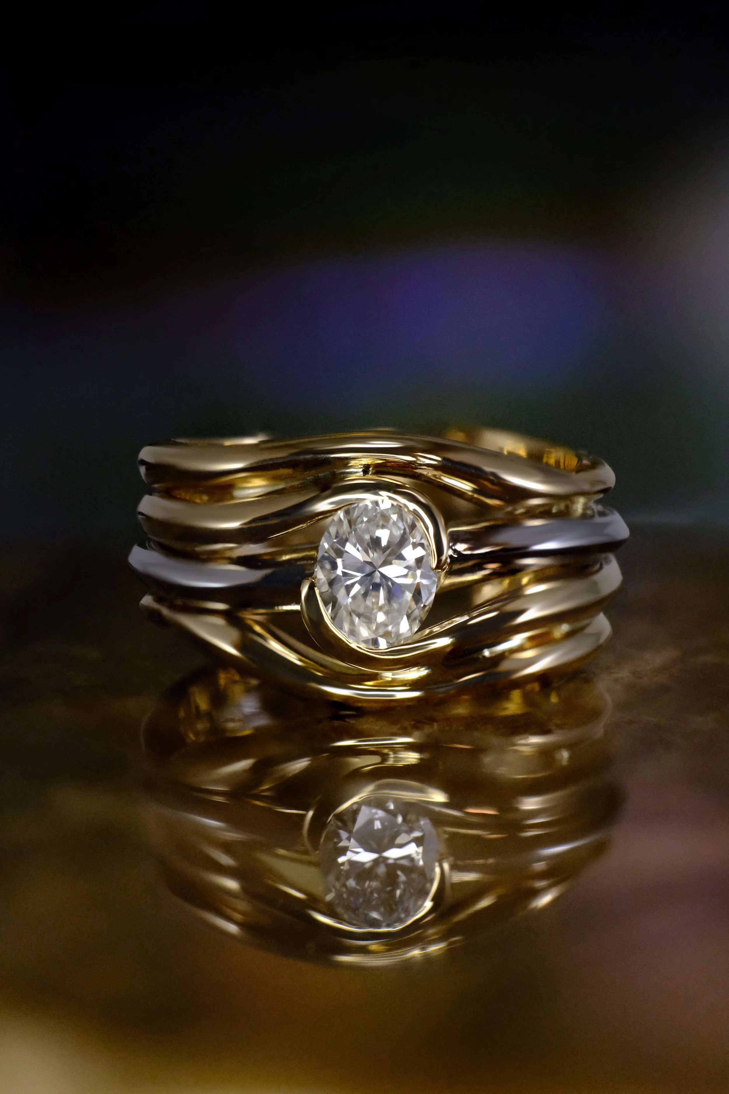 Front view of a two-tone ring combining white gold and yellow gold, featuring a round diamond set within flowing, layered bands that wrap around the stone, shown on a reflective surface with a dark, softly blurred background.