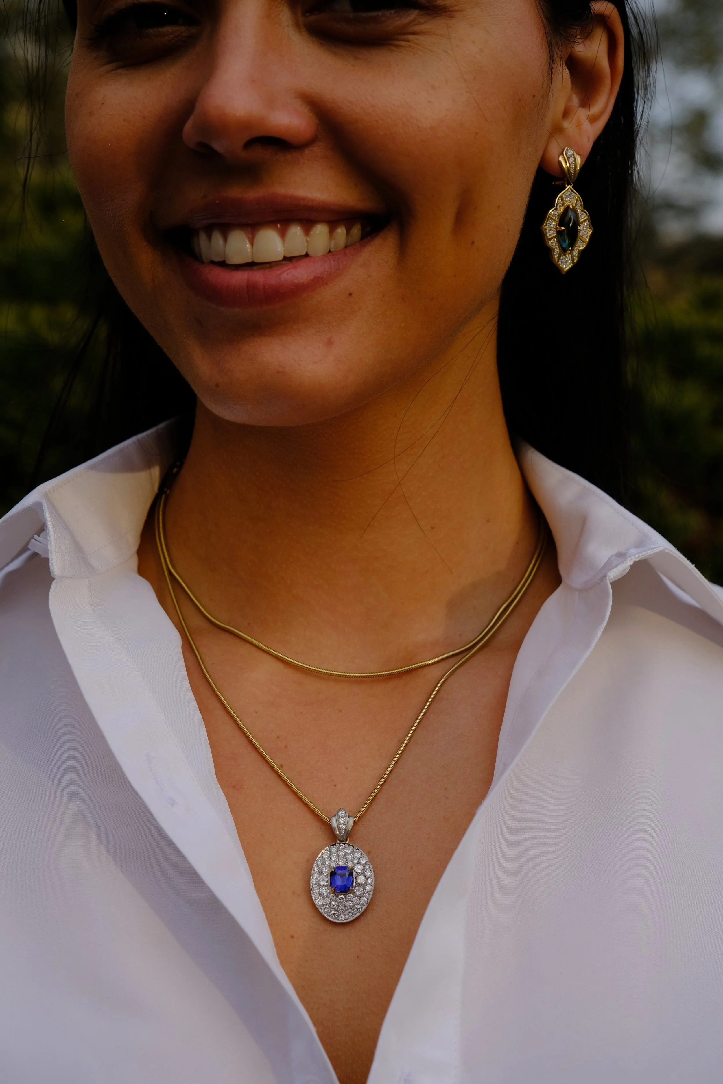 Gold chain necklace worn over a white blouse, featuring an oval pendant with a blue sapphire center surrounded by pavé diamonds, resting at the upper chest against a dark, blurred background.