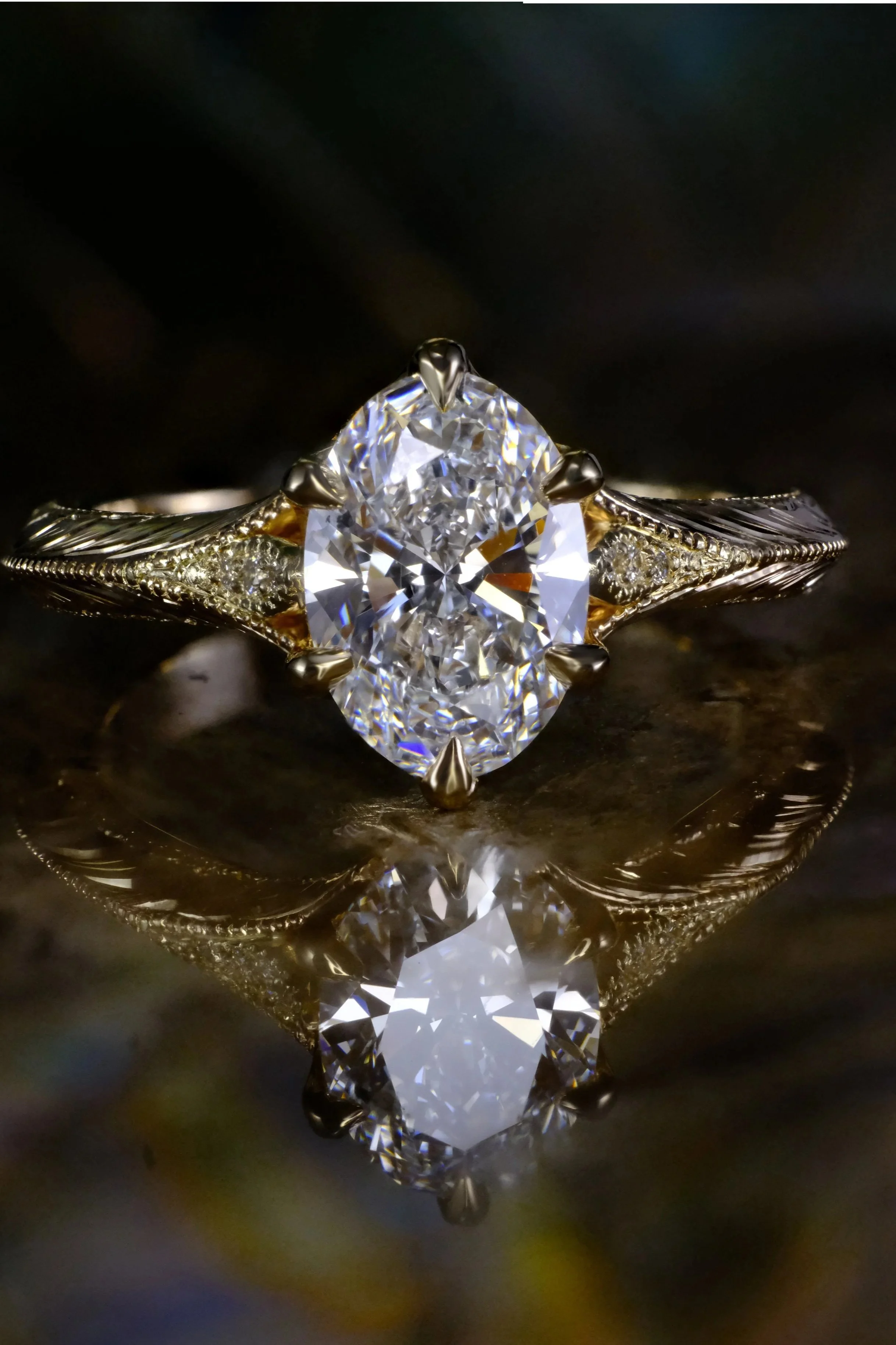 Front view of a yellow gold engagement ring with an oval diamond center, prong set, and engraved shoulders with small diamond accents.