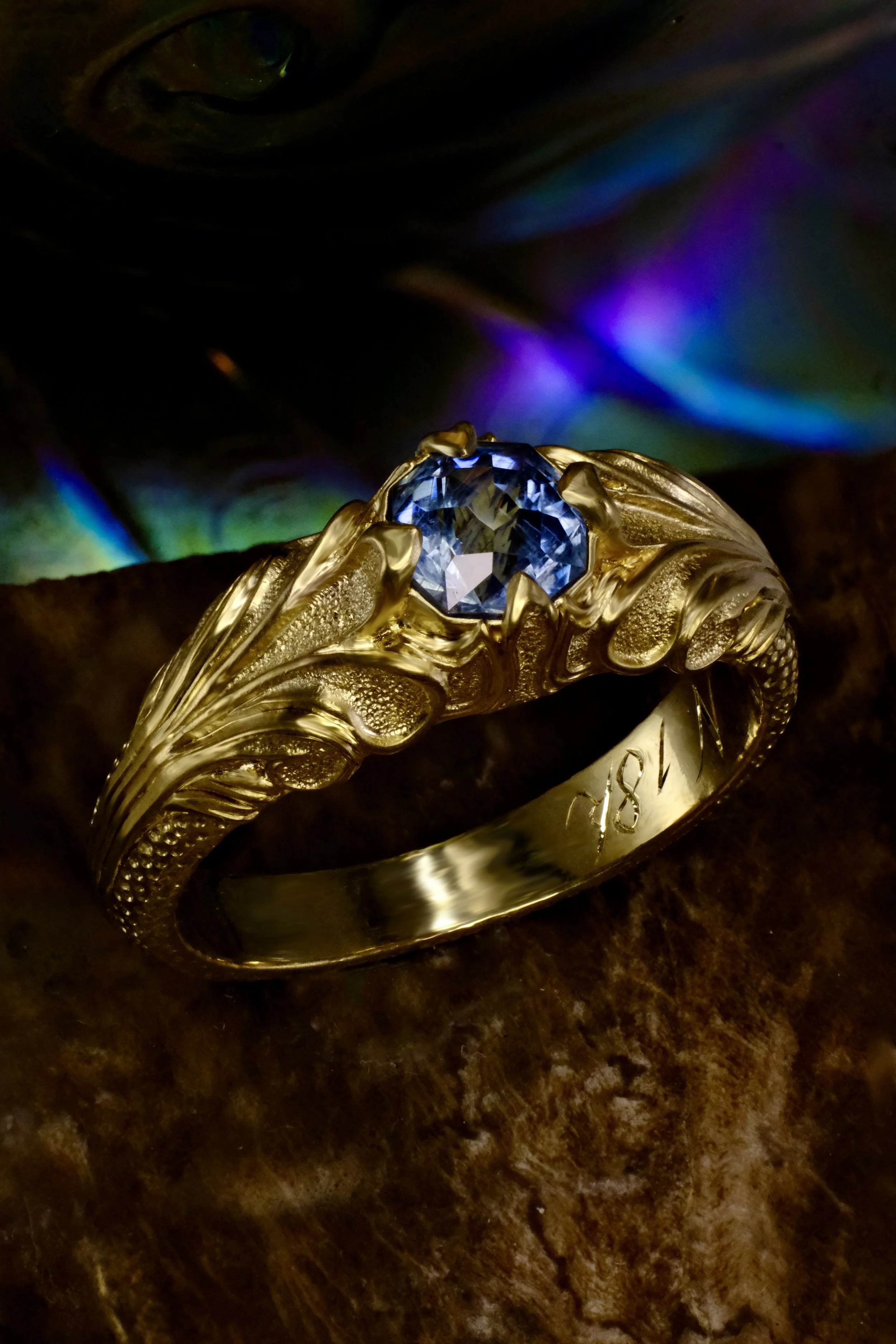 Yellow gold ring with blue sapphire in ornate leaf-engraved setting, shown straight on with reflection.