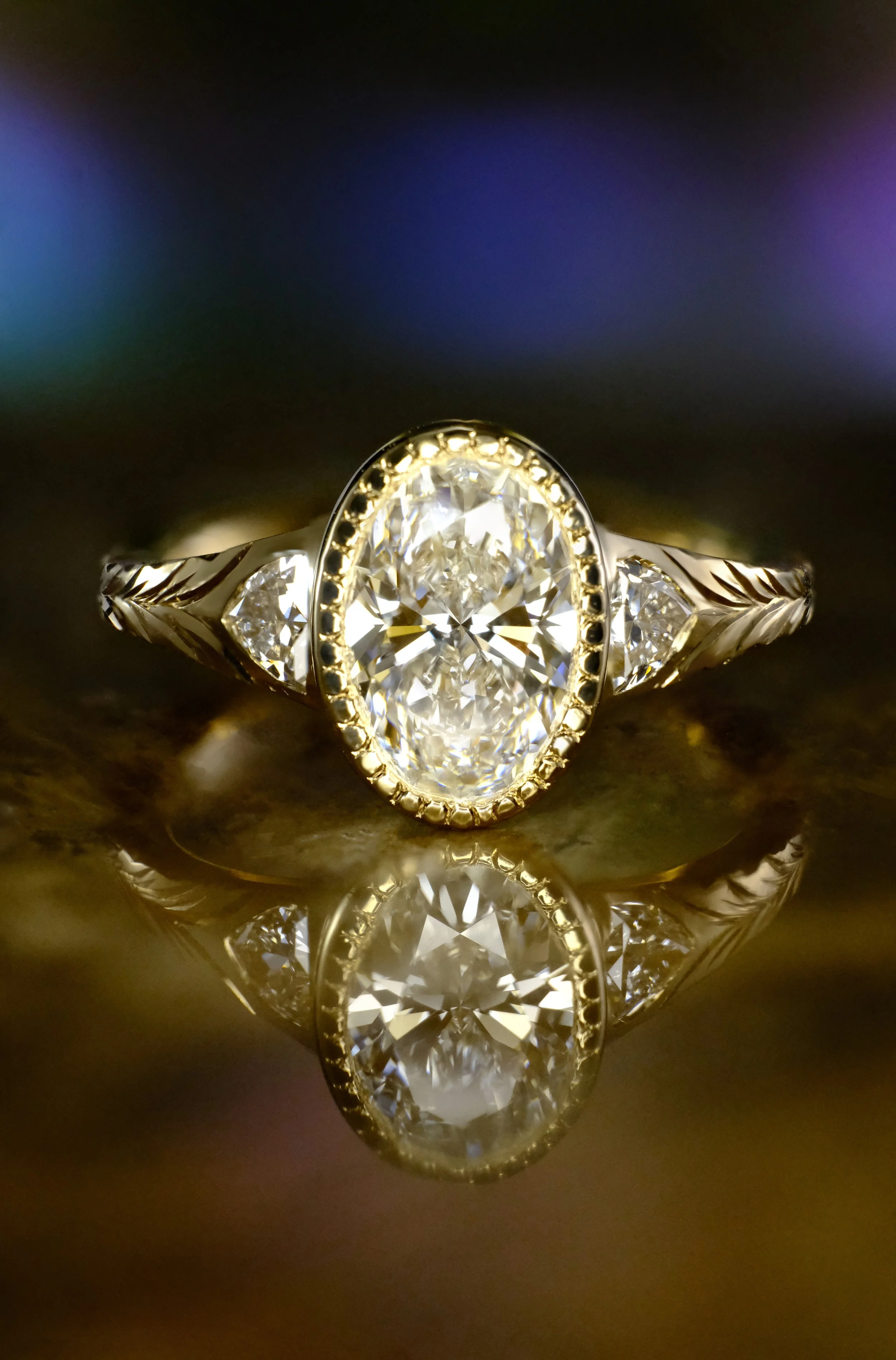 Yellow gold ring with oval diamond in bezel setting and side stones, shown head-on with reflection on a glossy surface.