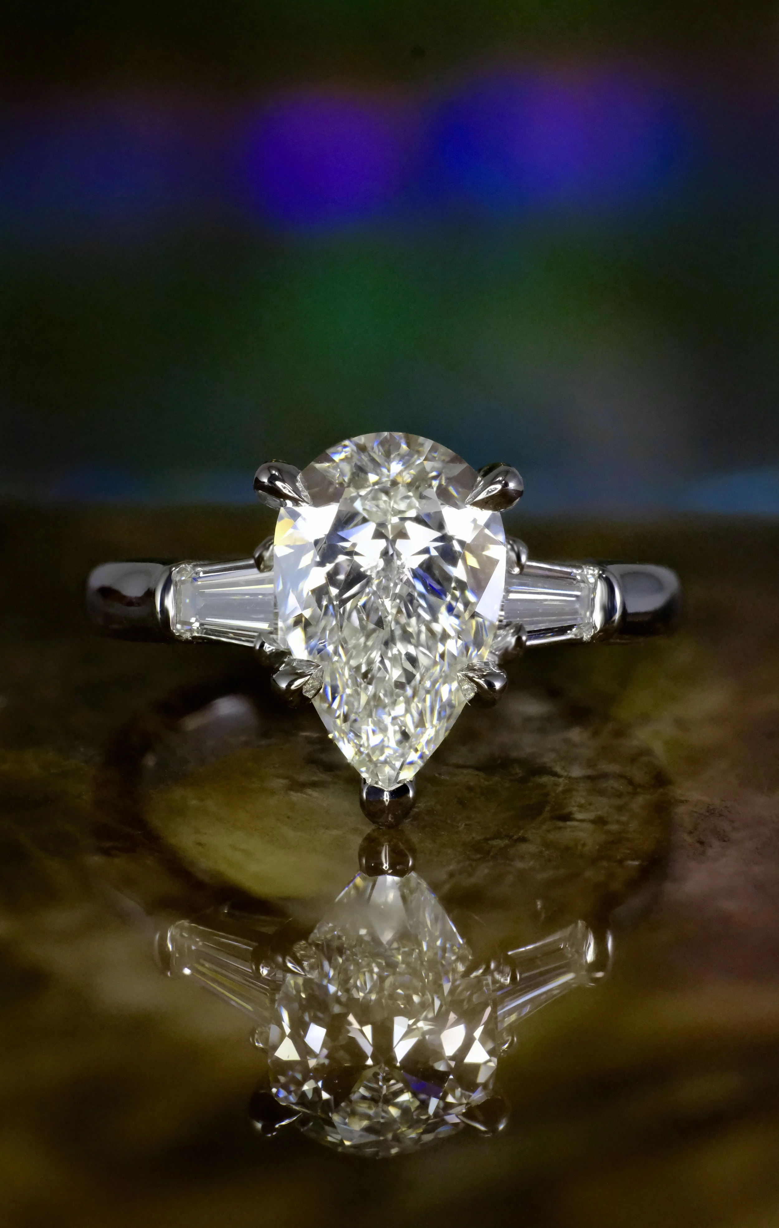 White gold ring with pear-shaped diamond and tapered baguette side stones, shown head-on with reflection.