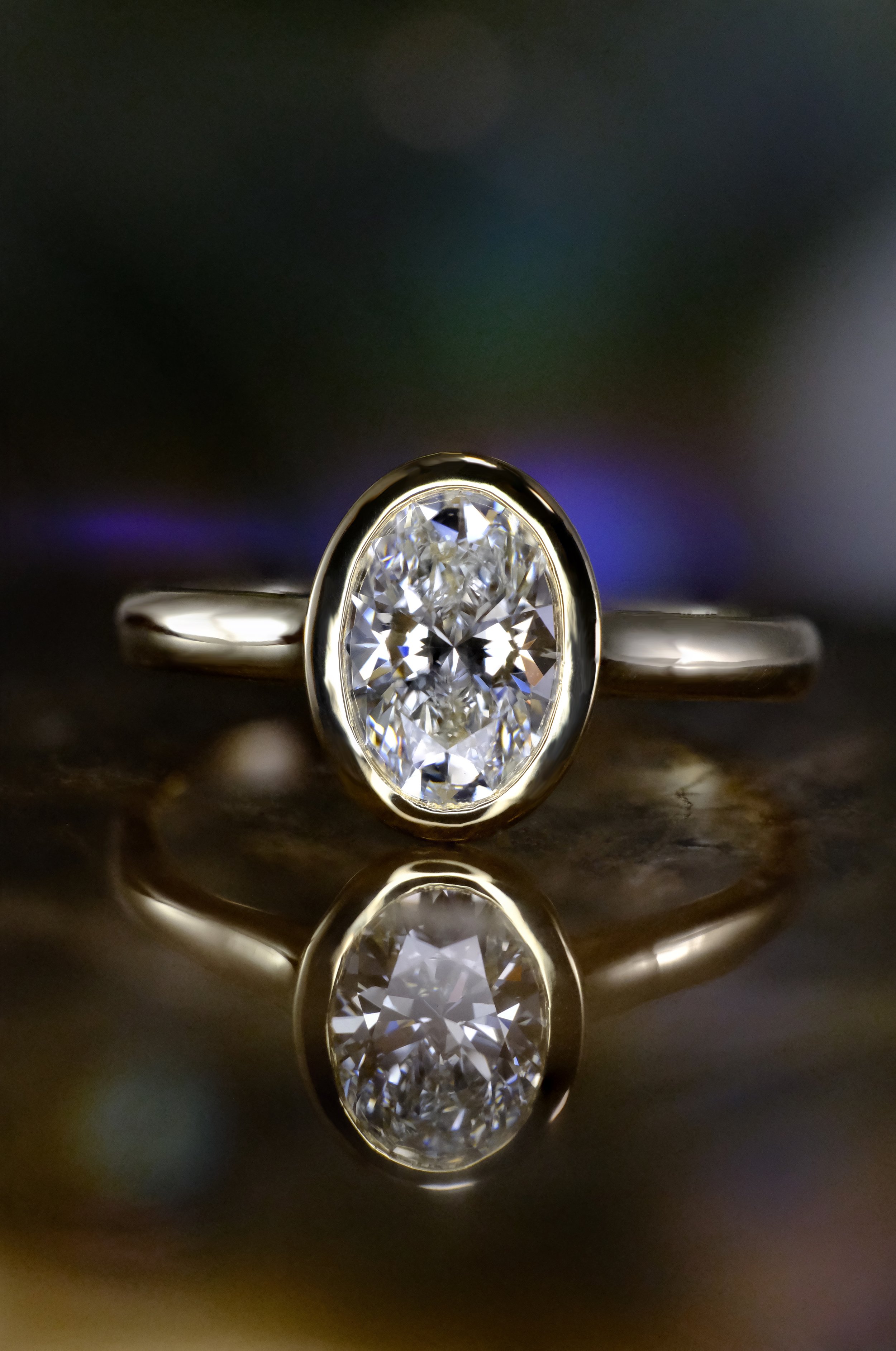 White metal (white gold or platinum) ring with an oval diamond in a bezel setting, shown head-on with the centered stone and its reflection visible below on a polished surface.