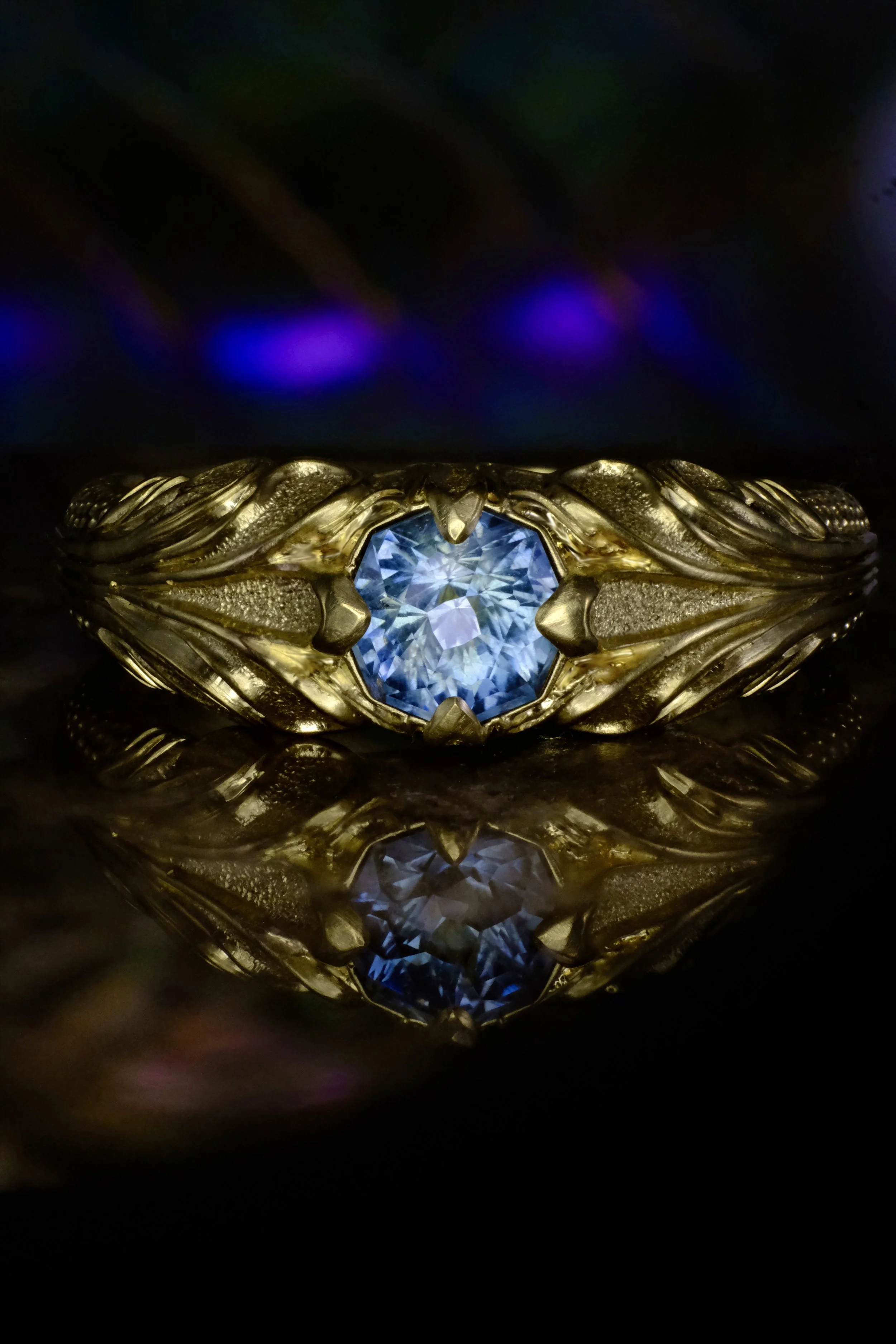 Yellow gold ring with blue sapphire in ornate leaf-engraved setting, shown straight on with reflection.